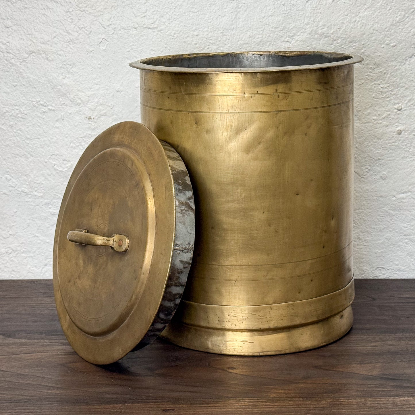 Brass trash can with lid on a wooden surface against a white wall