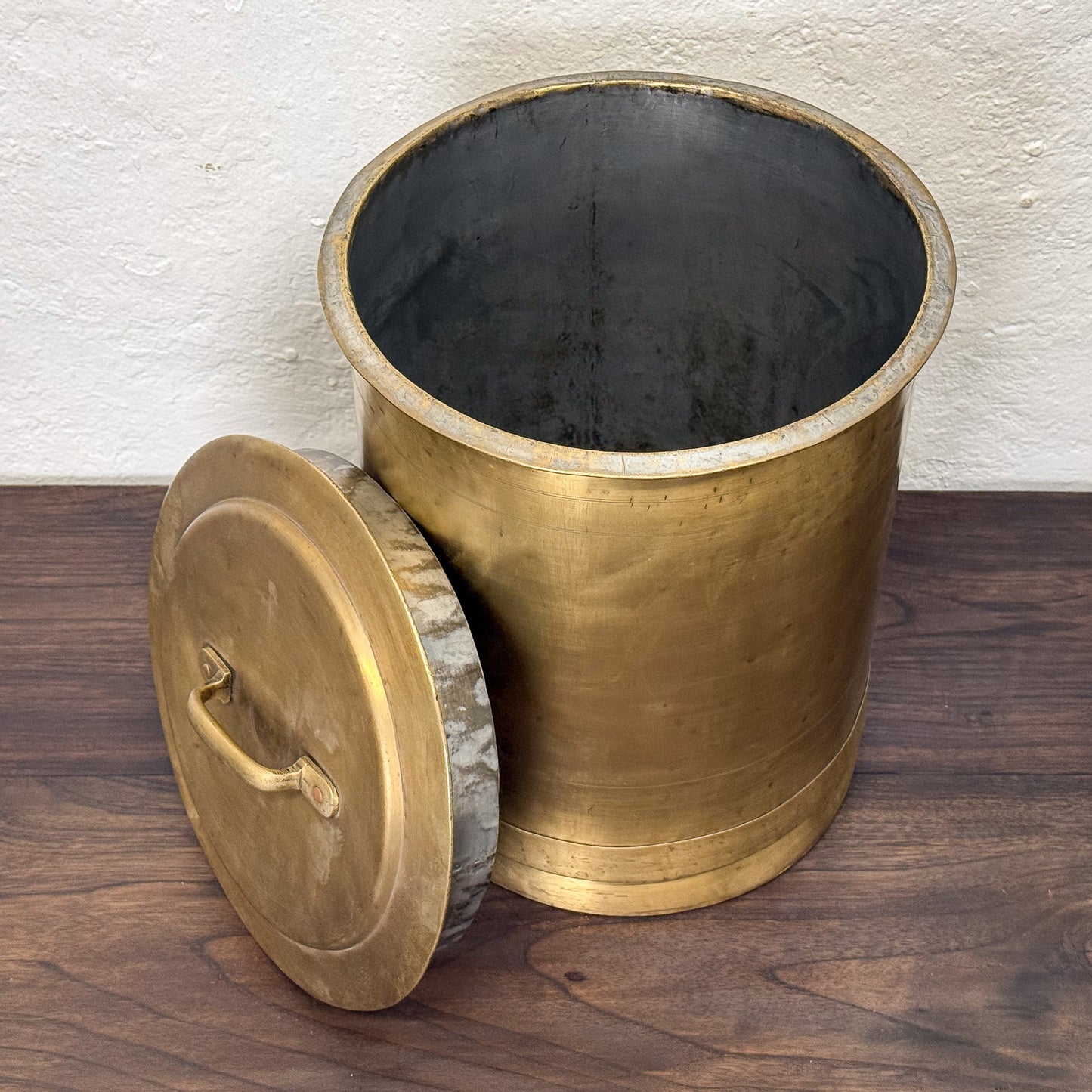 Brass trash bin with lid on a wooden floor against a white wall