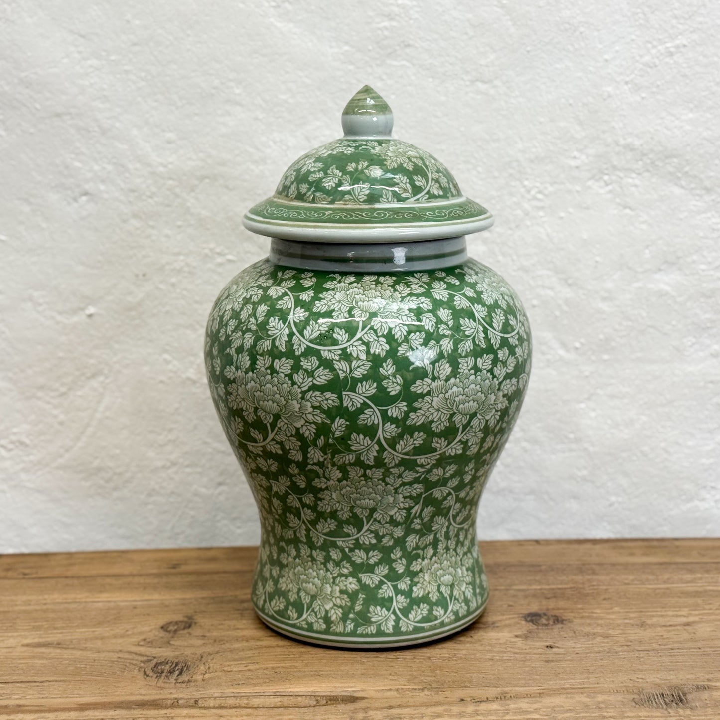 Green ceramic jar with floral patterns on a wooden surface and white textured wall background