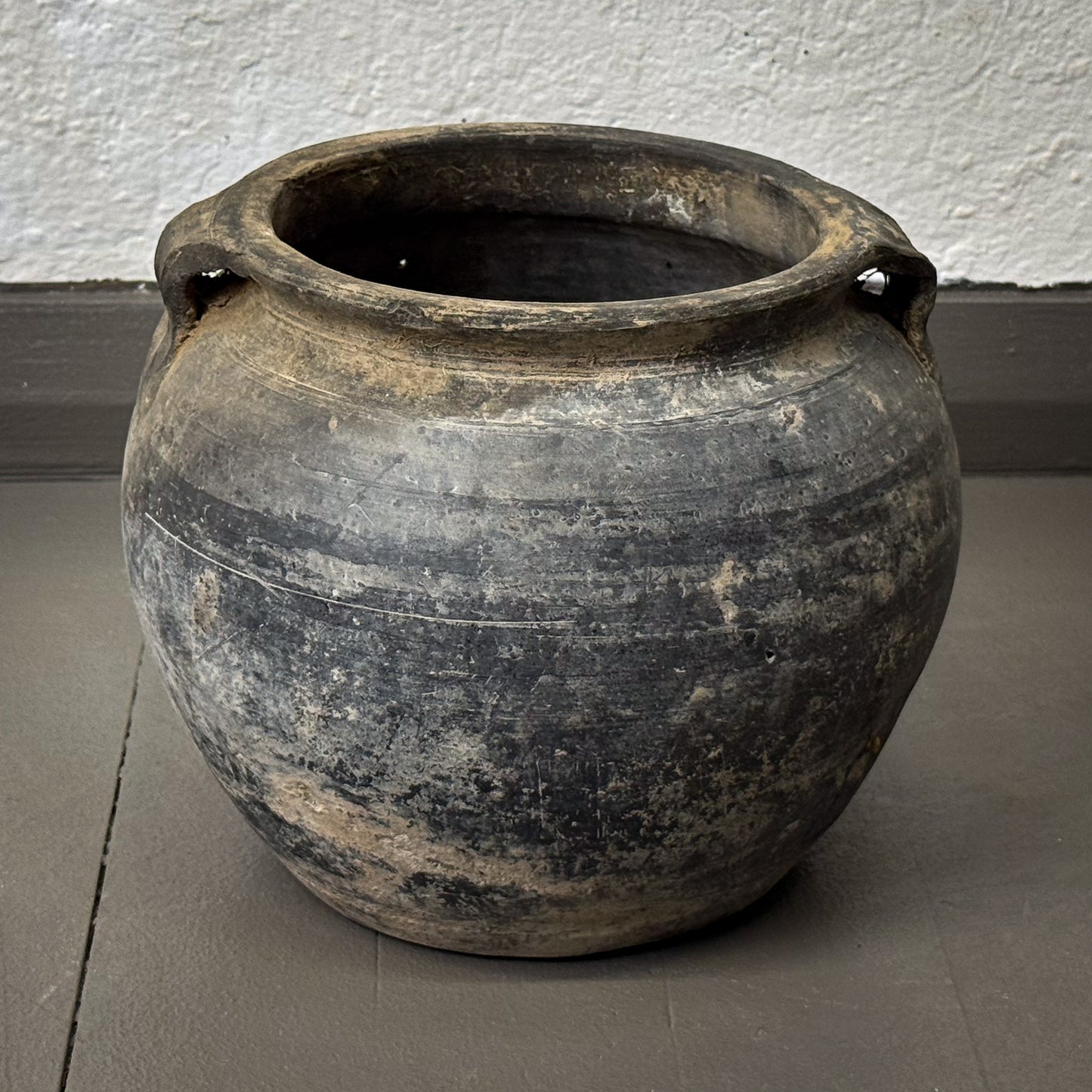 Vintage terracotta pot with handles on a gray floor against a white wall.
