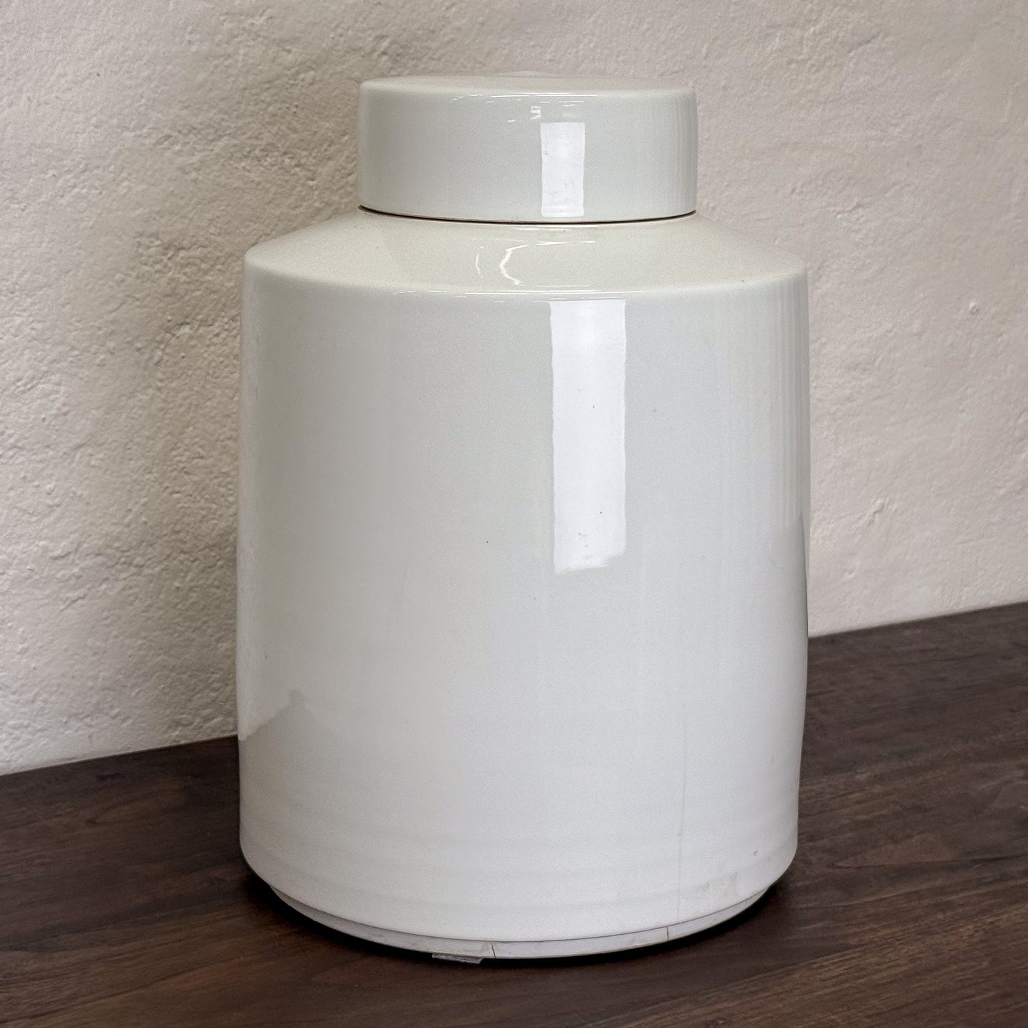 White ceramic jar with a lid on a wooden surface against a textured wall.