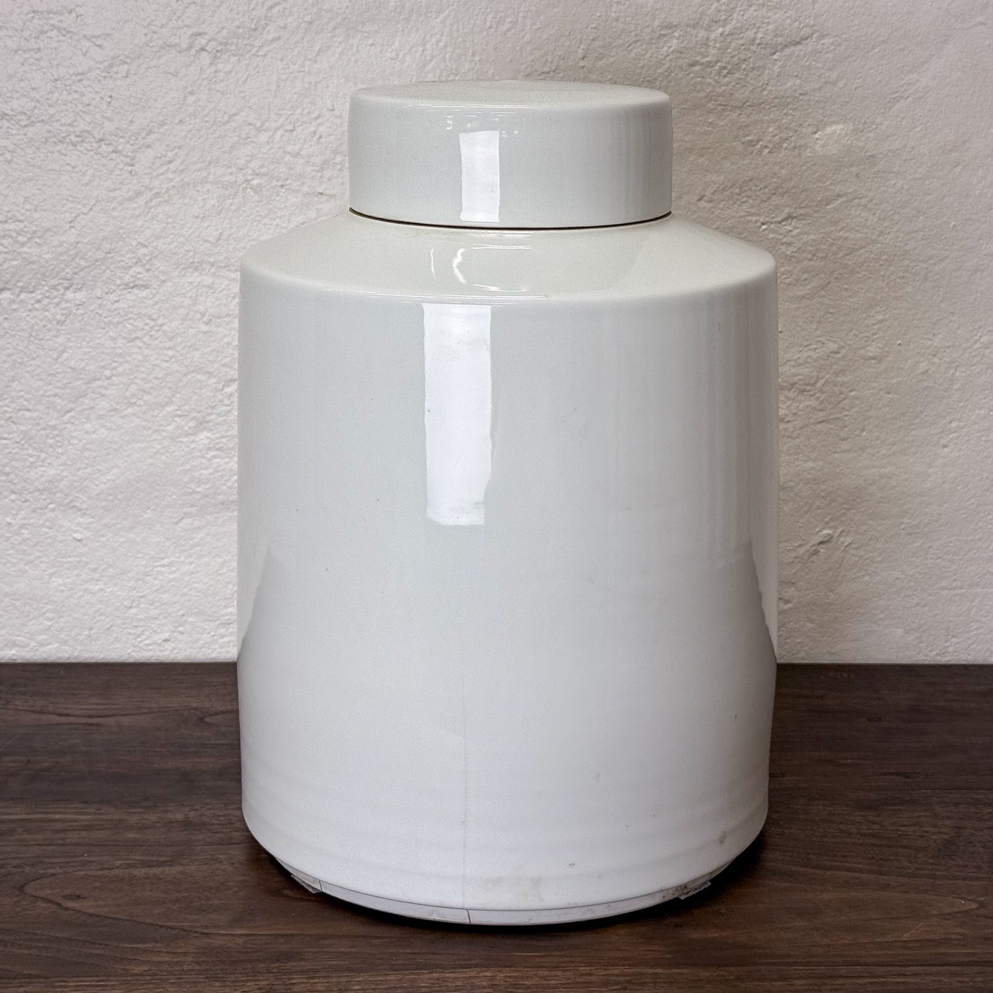 White ceramic jar with a lid on a wooden surface against a textured wall.