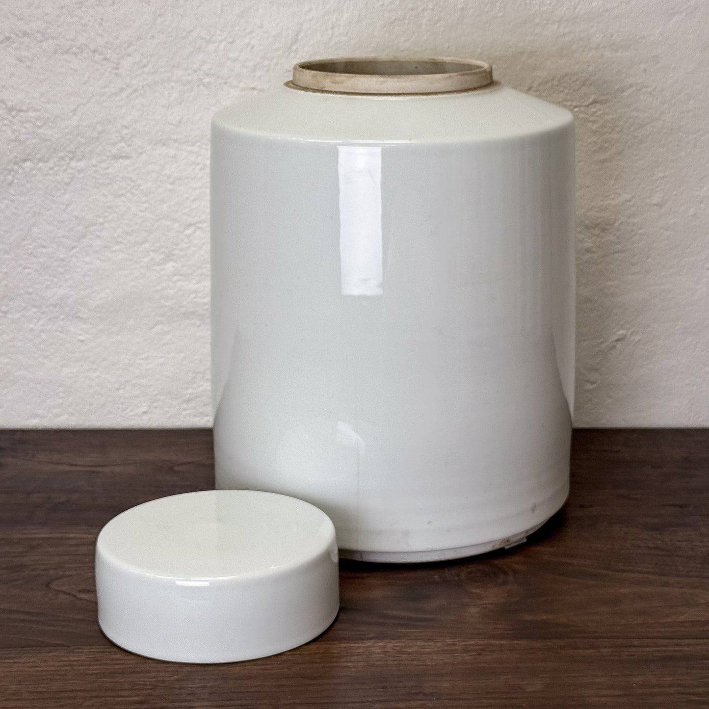 Two white ceramic jars with lids on a wooden surface against a textured wall.