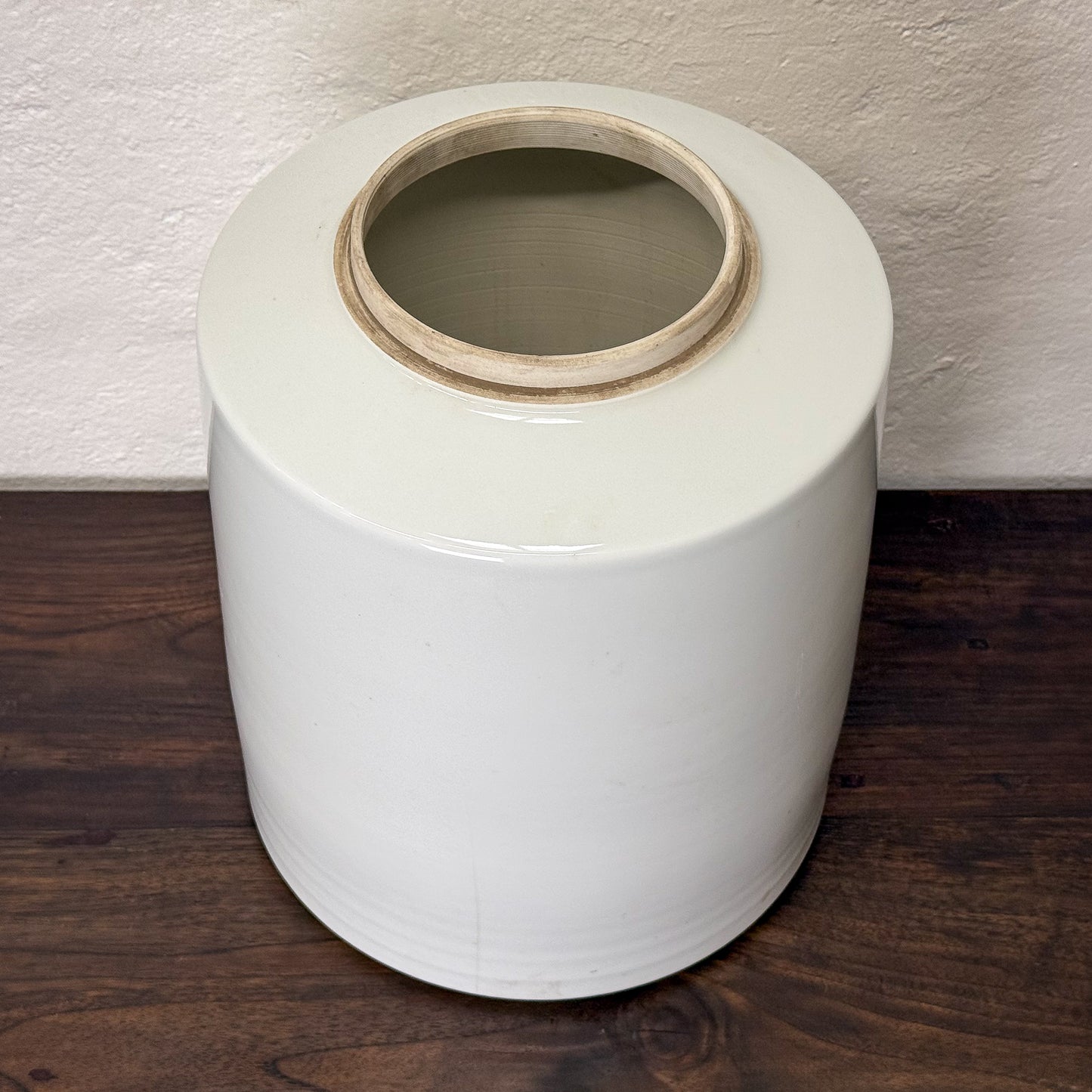 White ceramic jar with a lid on a wooden surface against a textured wall.