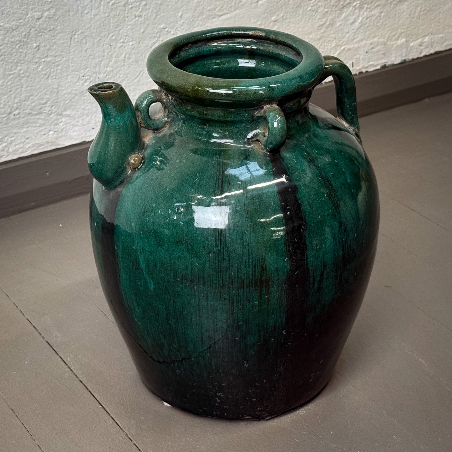 Green ceramic jar with handles on a gray floor against a white wall.