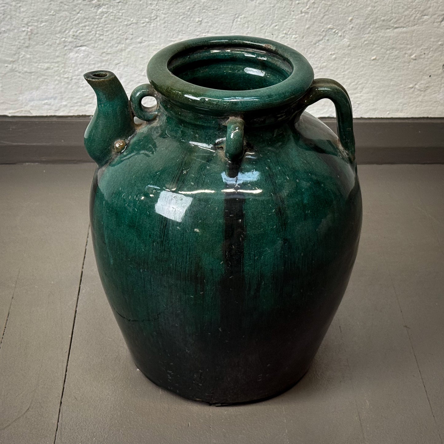 Green ceramic jar with handles on a gray floor against a white wall.