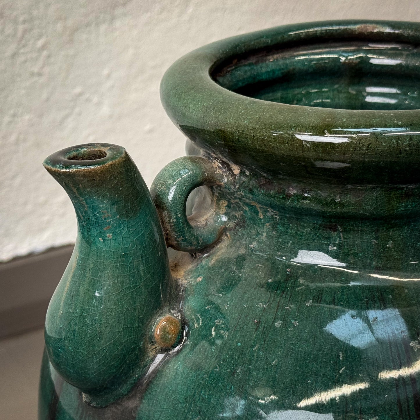 Green ceramic pot with a spout against a textured wall.