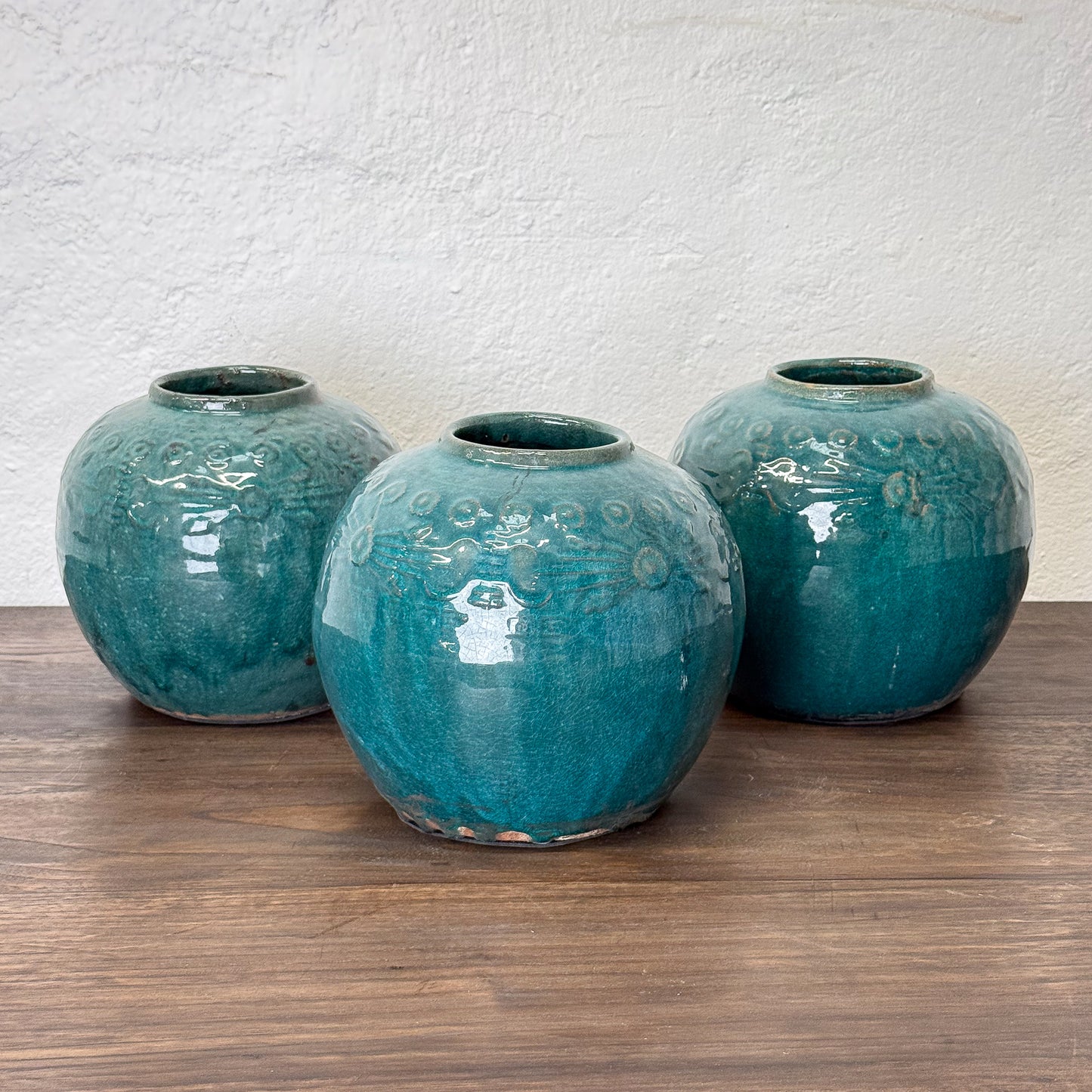 Three teal ceramic vases on a wooden surface with a white textured wall background