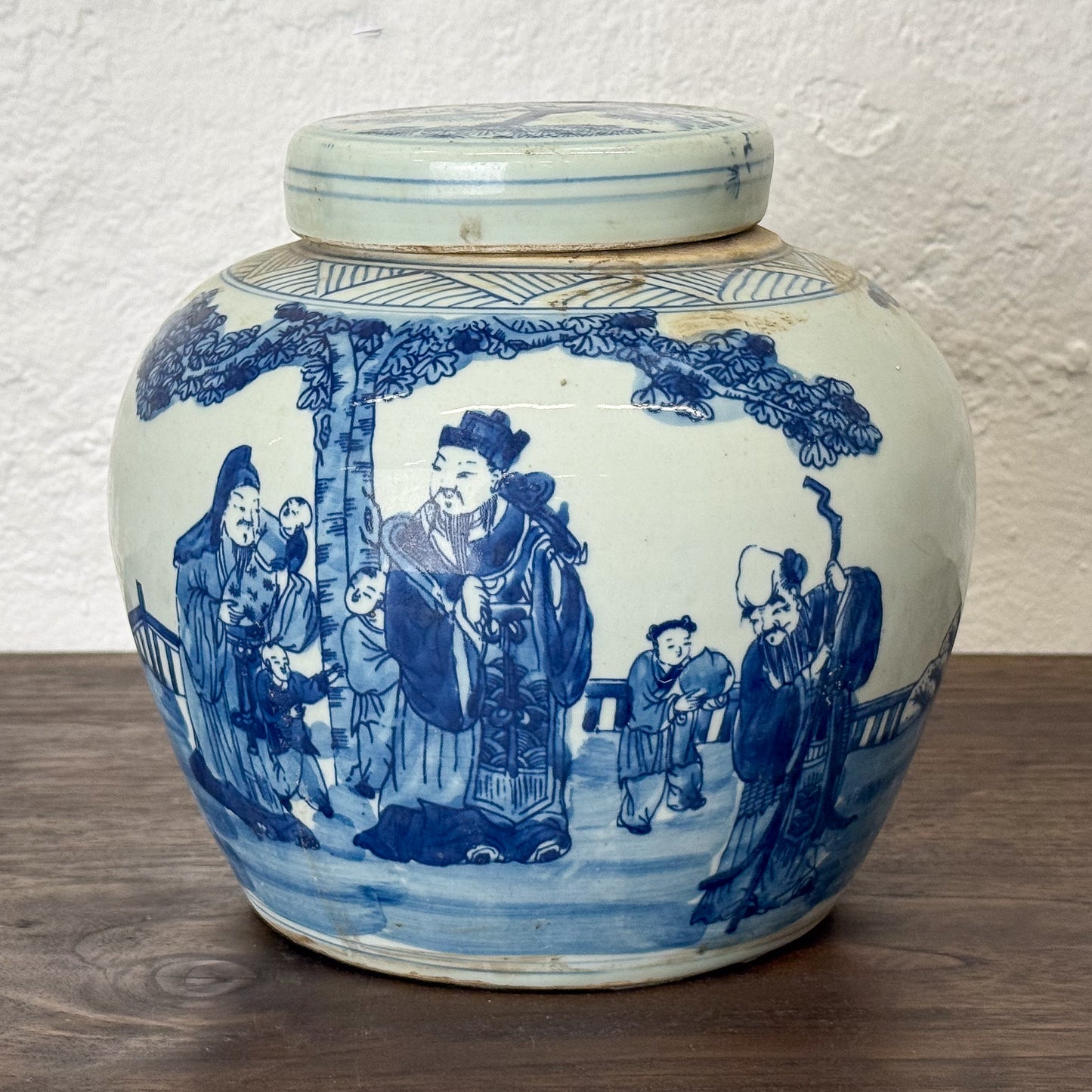 Blue and white ceramic jar with lid featuring classical Chinese scenes on a wooden surface.