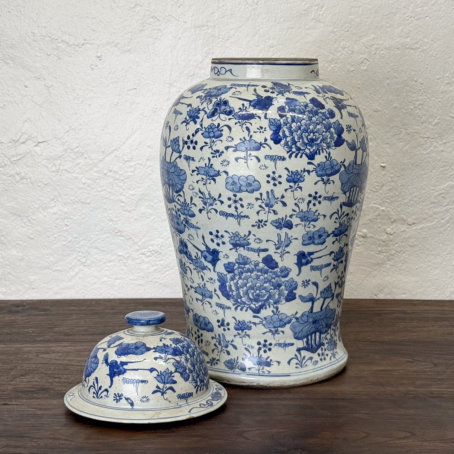 Blue and white ceramic jar with matching cover on a wooden surface against a textured white wall.