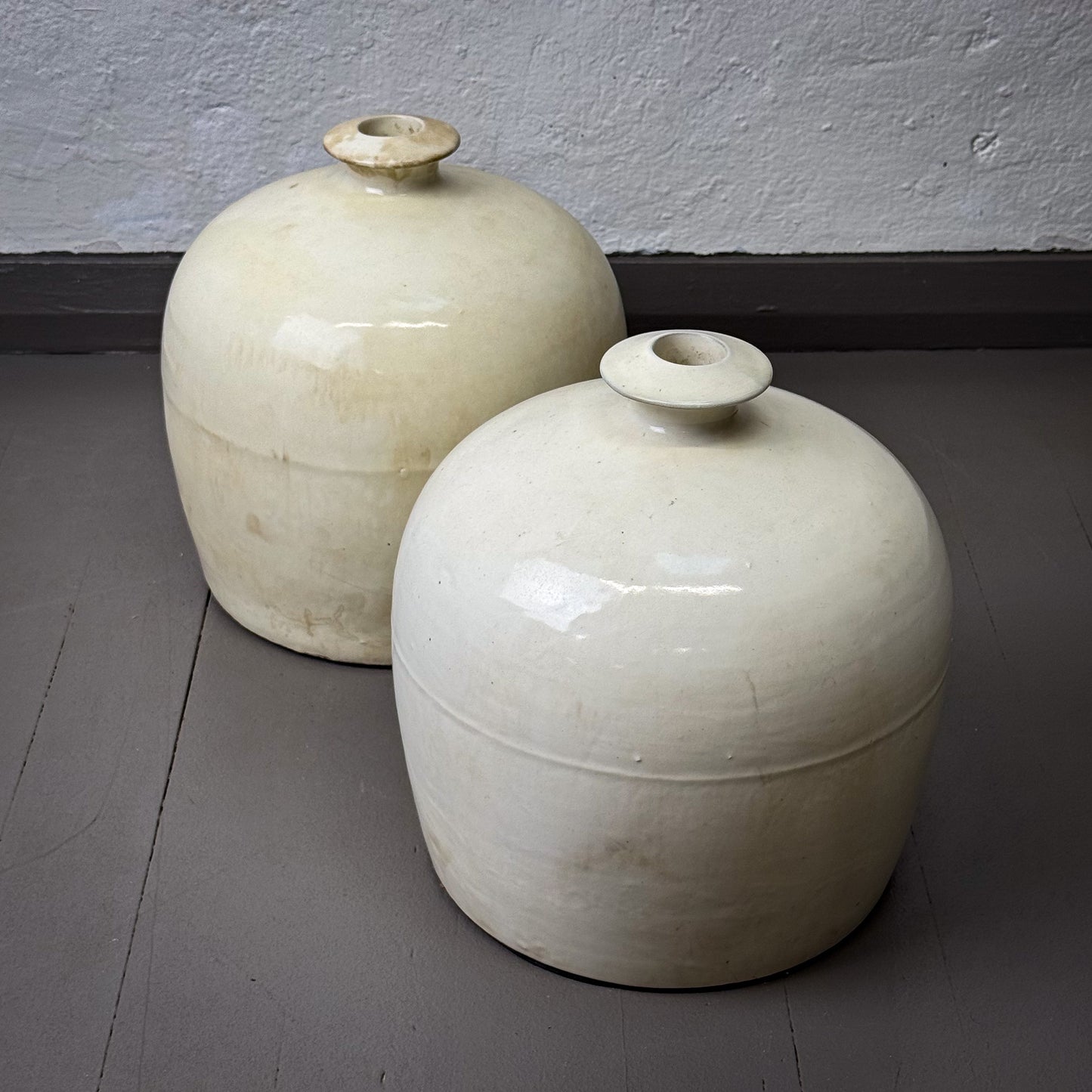 Two white ceramic jars with lids on a gray floor against a plain wall.