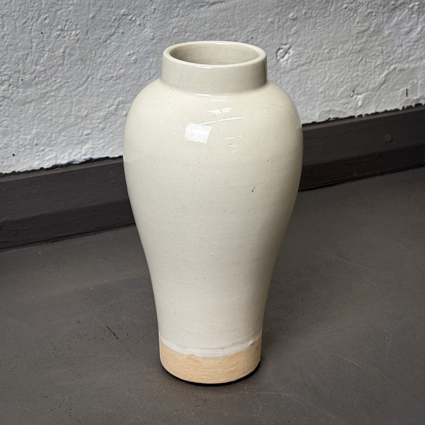 White ceramic vase with a wooden base on a gray floor against a white wall.