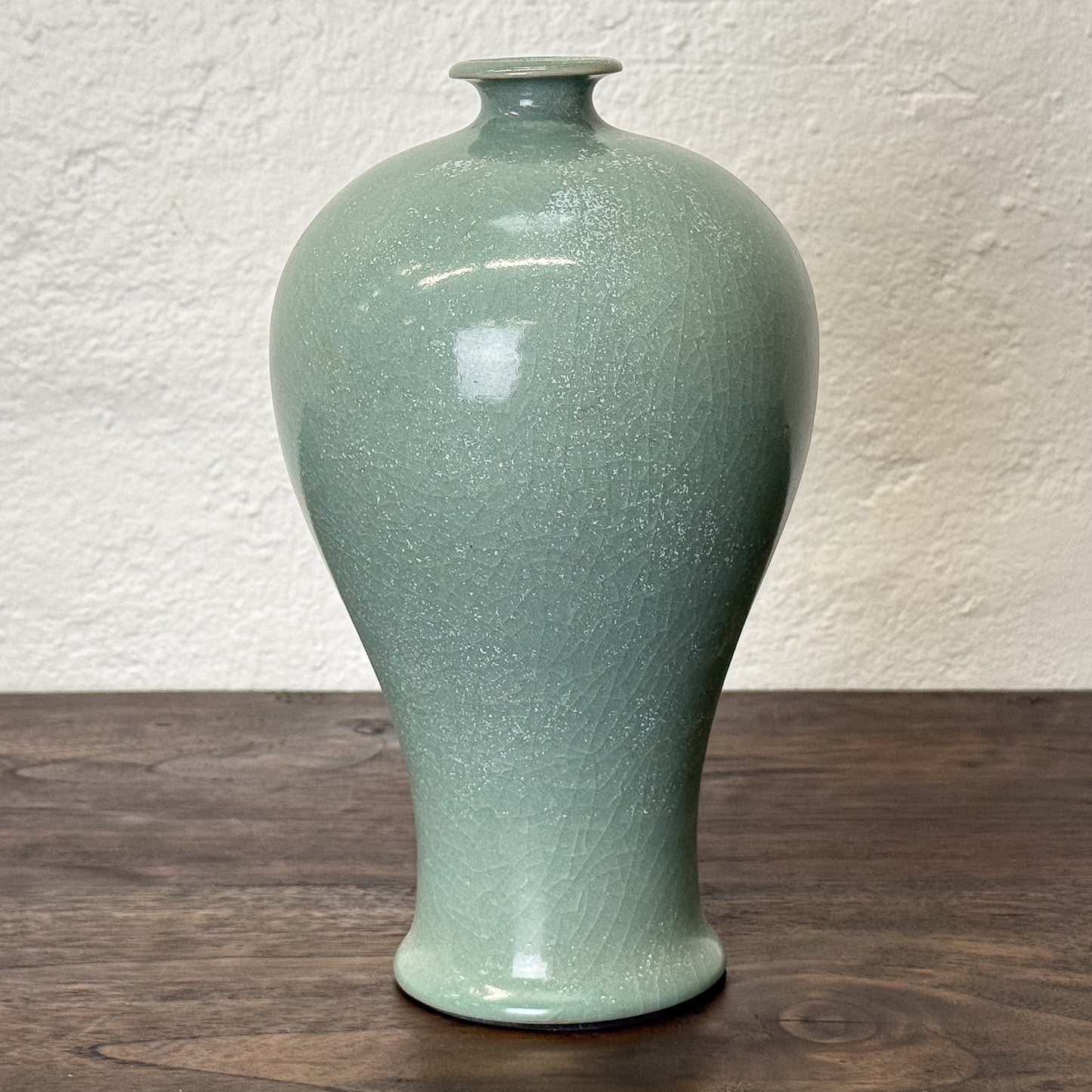 Green ceramic vase on a wooden surface with a white textured wall background