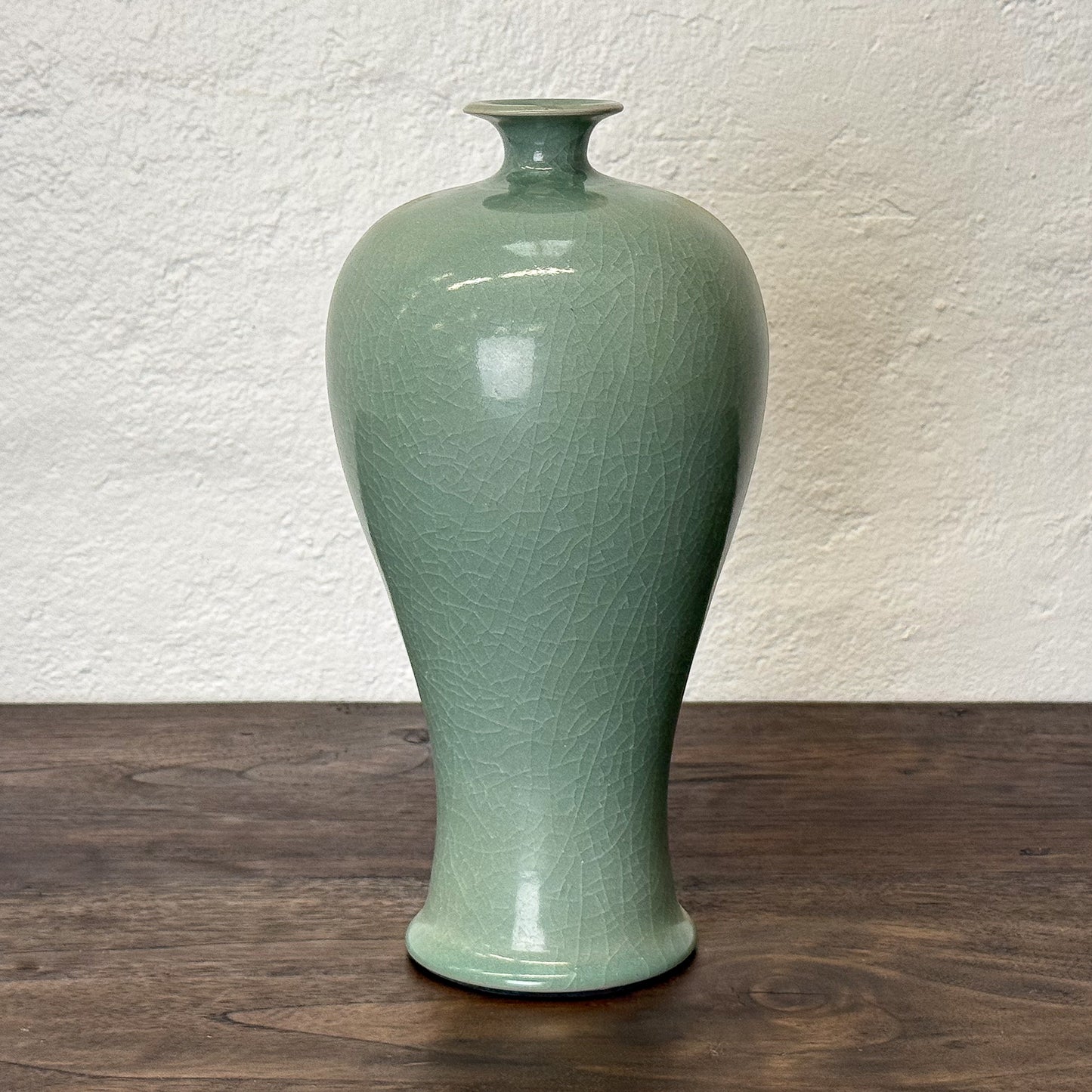 Green ceramic vase on a wooden surface with a textured white wall background