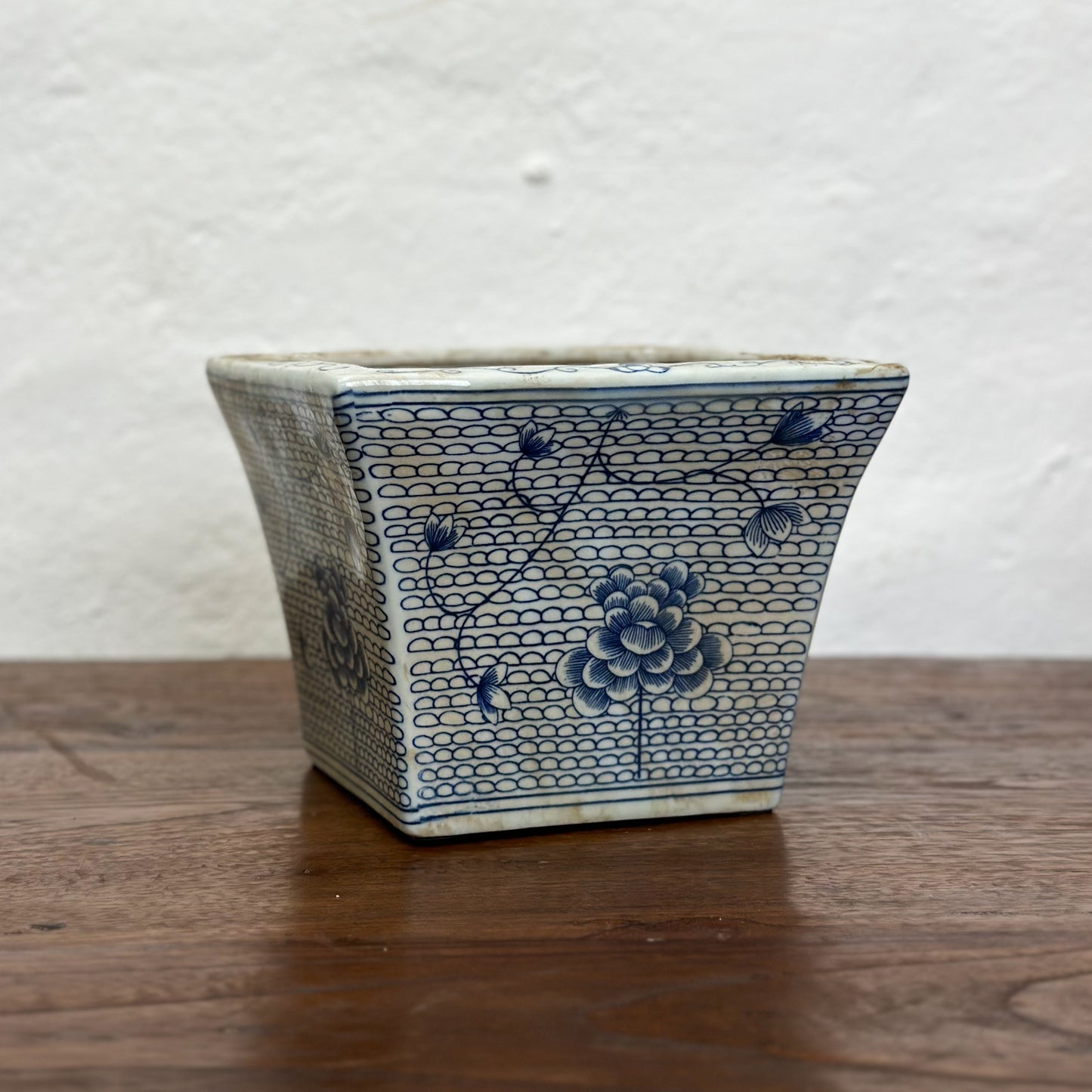Two square porcelain planters with a blue floral pattern on a beige background, displayed on a wooden surface against a plain wall.