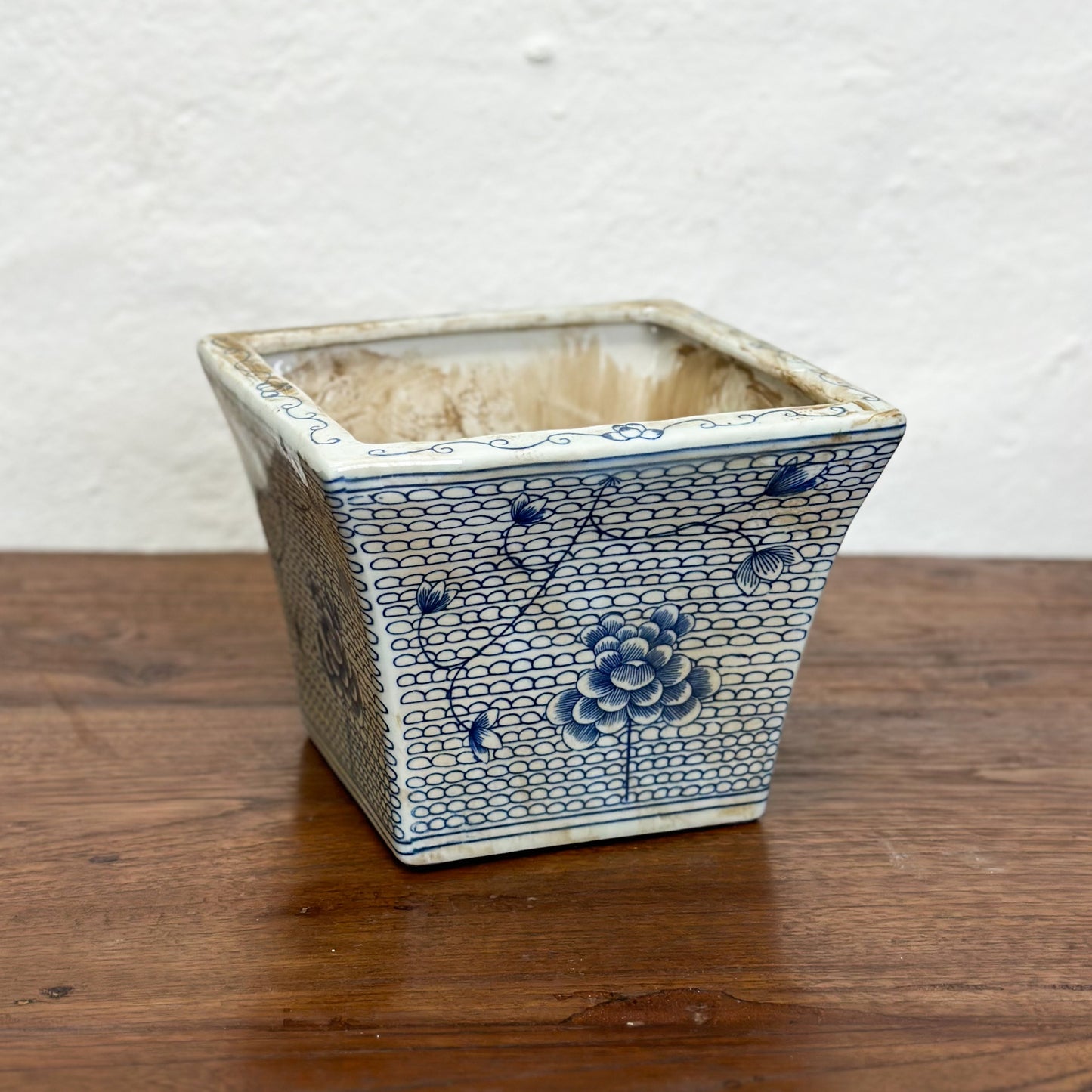 Two square porcelain planters with a blue floral pattern on a beige background, displayed on a wooden surface against a plain wall.
