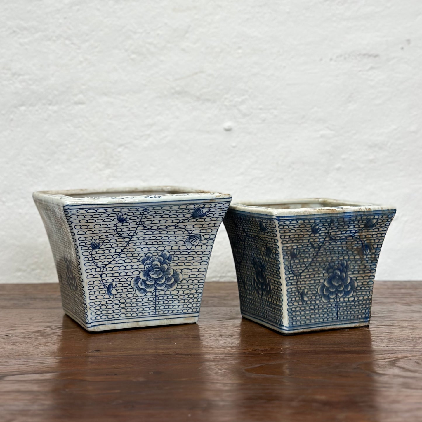 Two square porcelain planters with a blue floral pattern on a beige background, displayed on a wooden surface against a plain wall.