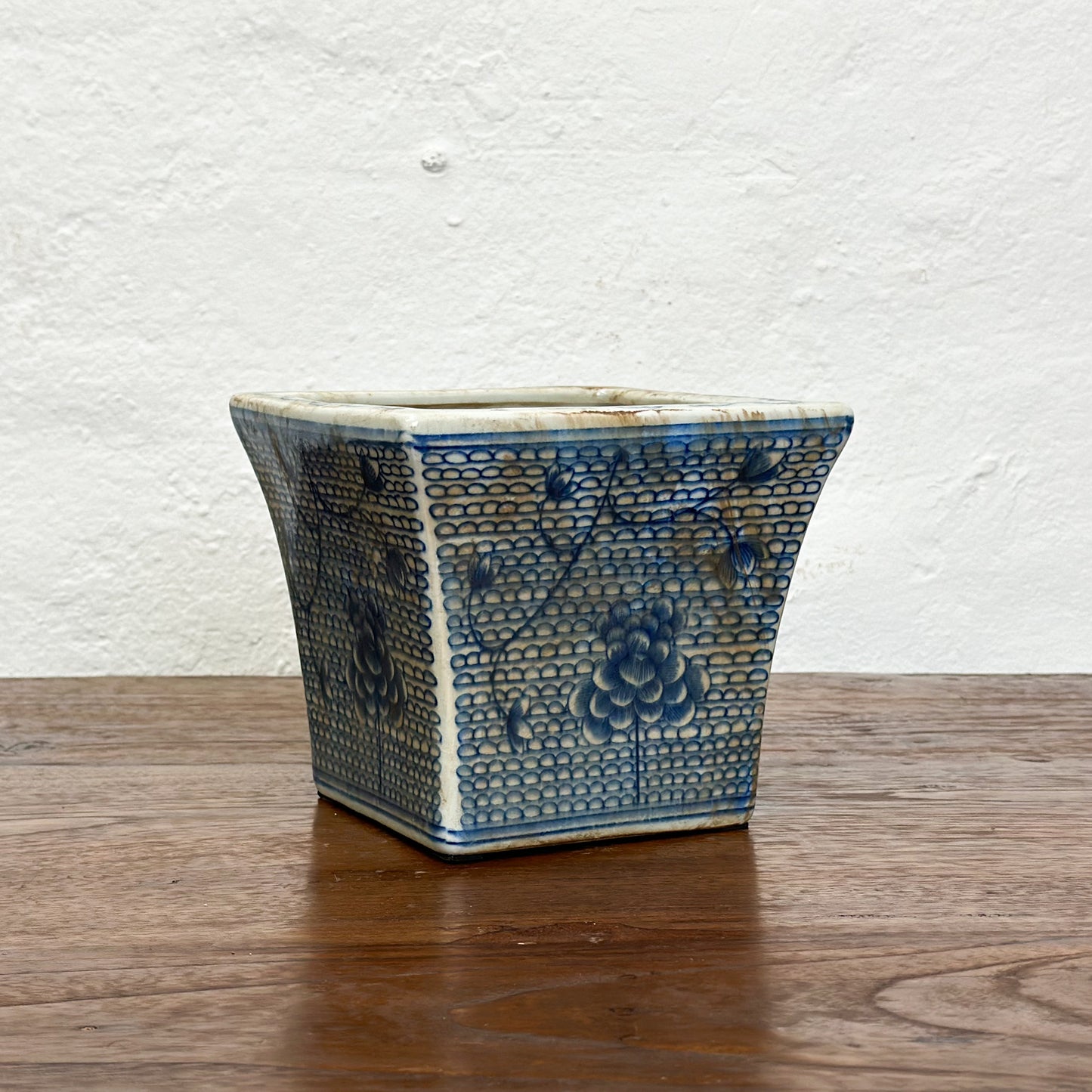 Two square porcelain planters with a blue floral pattern on a beige background, displayed on a wooden surface against a plain wall.