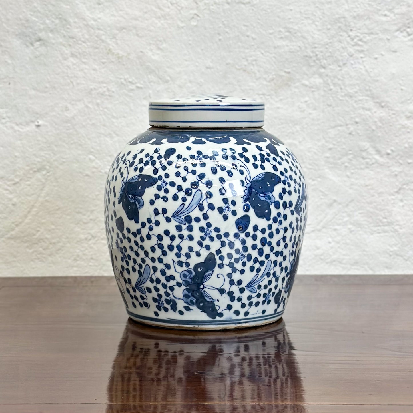 A blue porcelain ginger jar with a butterfly pattern and dotted details, displayed on a wooden surface against a textured background.