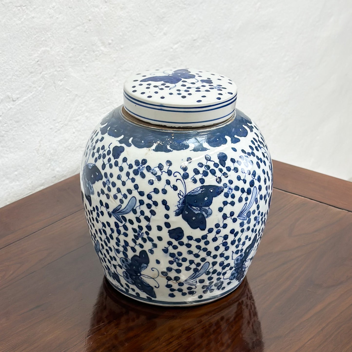 A blue porcelain ginger jar with a butterfly pattern and dotted details, displayed on a wooden surface against a textured background.