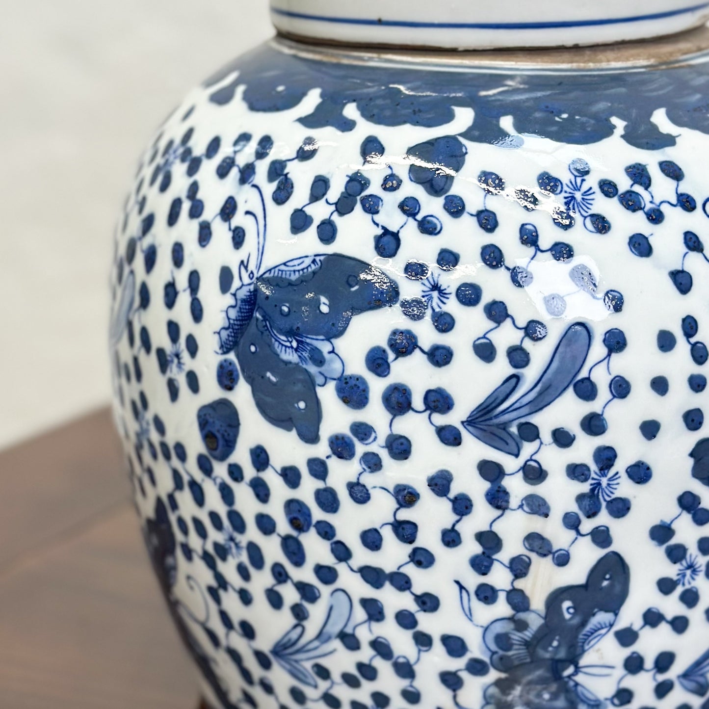 A blue porcelain ginger jar with a butterfly pattern and dotted details, displayed on a wooden surface against a textured background.