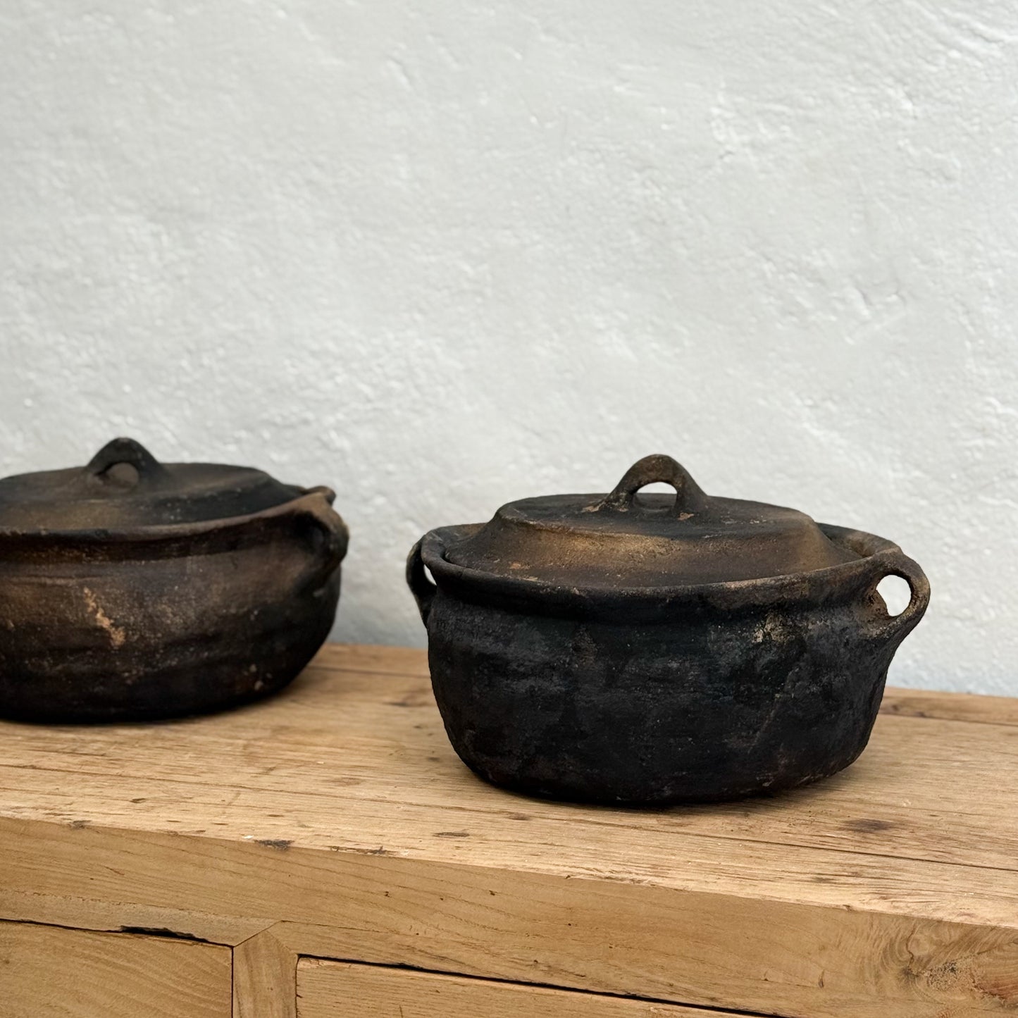 Two blackened iron pots with lids on a wooden surface against a textured white wall.