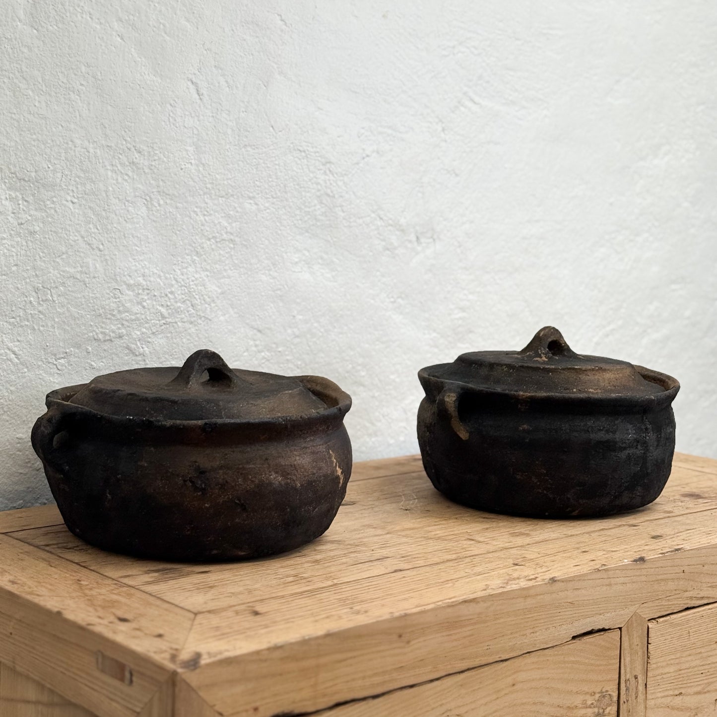 Two black ceramic pots with lids on a wooden surface against a white wall.