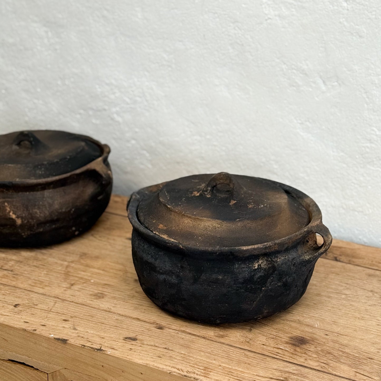 Two old, blackened pots with lids on a wooden surface against a white wall.
