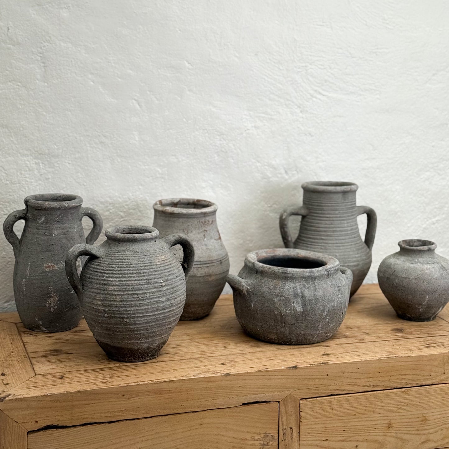 Set of six rustic gray clay pots on a wooden surface with a white textured wall background.