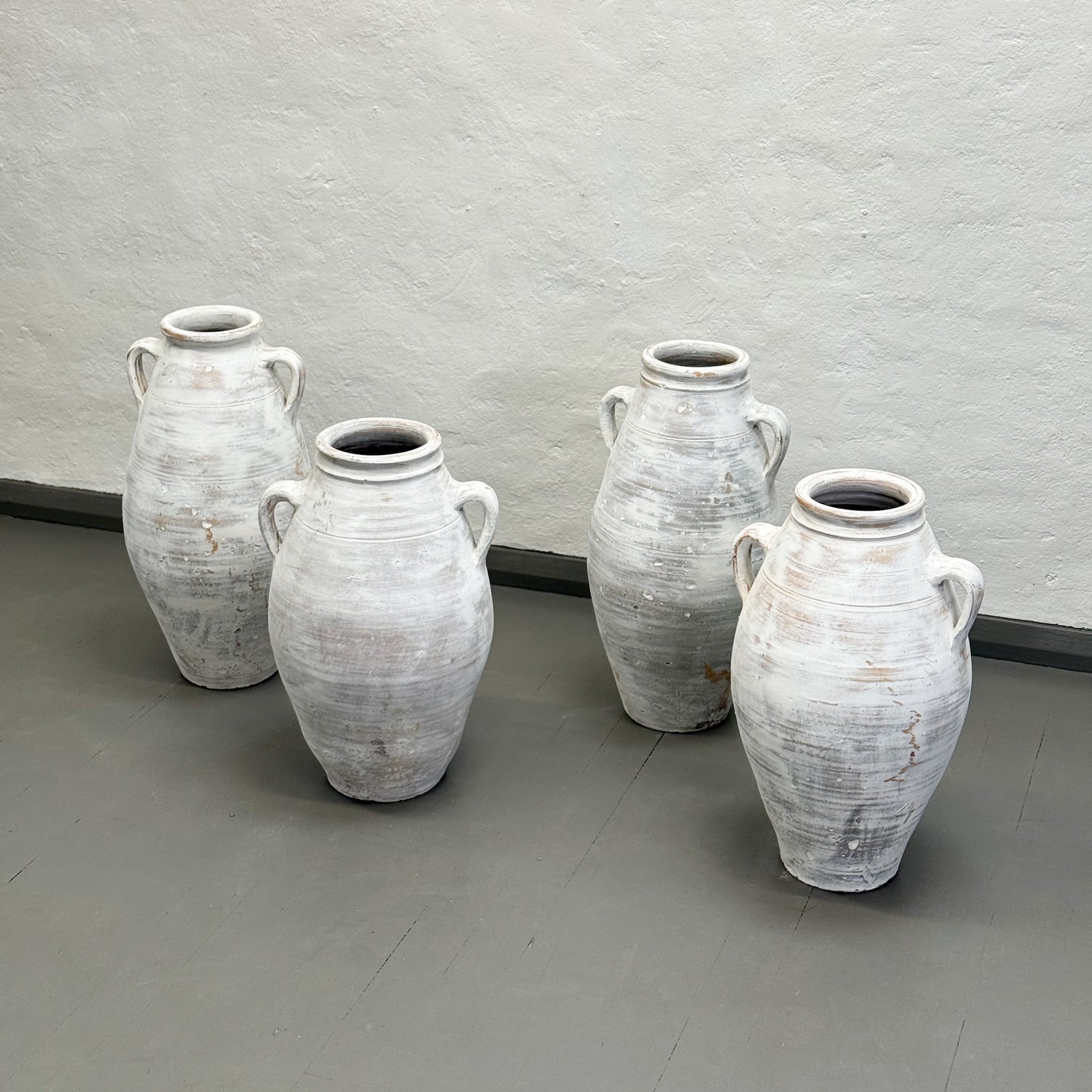Four white ceramic vases with handles on a gray floor against a gray wall.