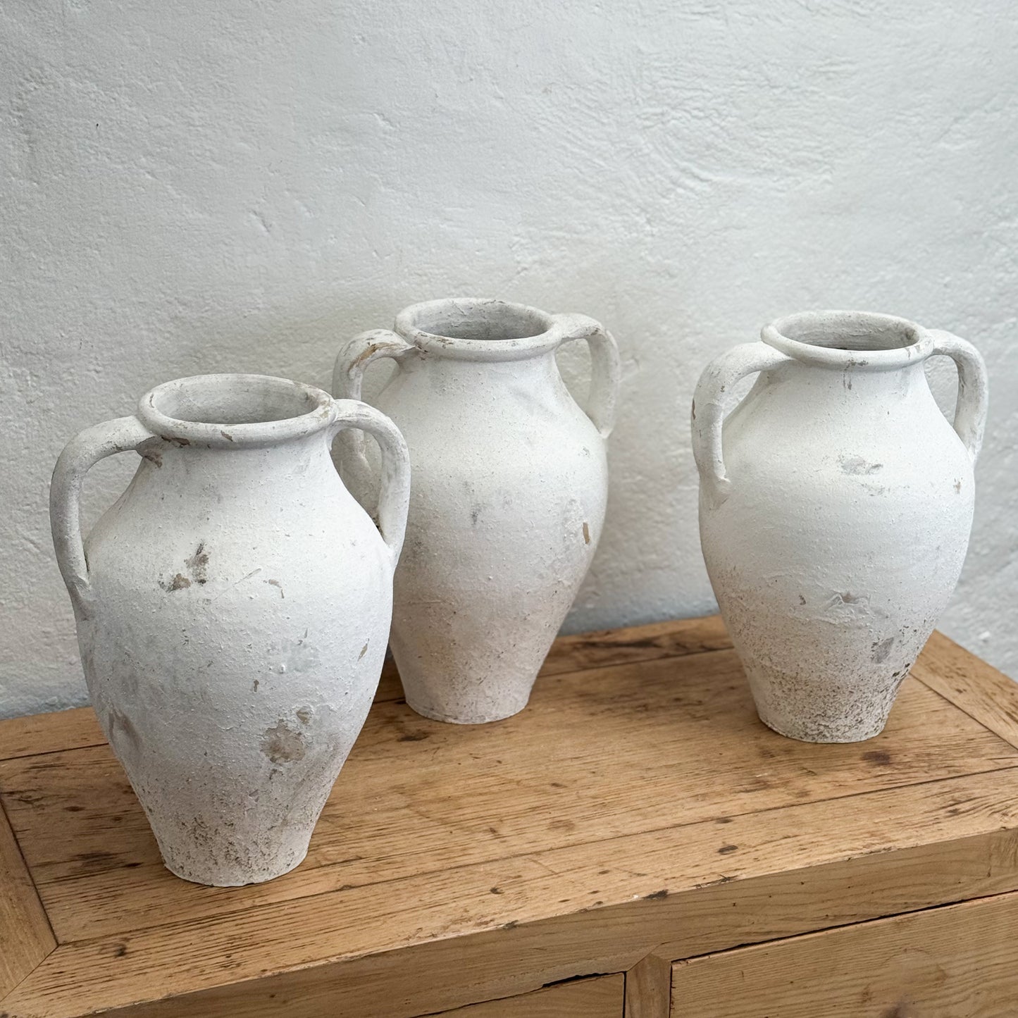 Three rustic white ceramic vases on a wooden surface with a light gray wall background.