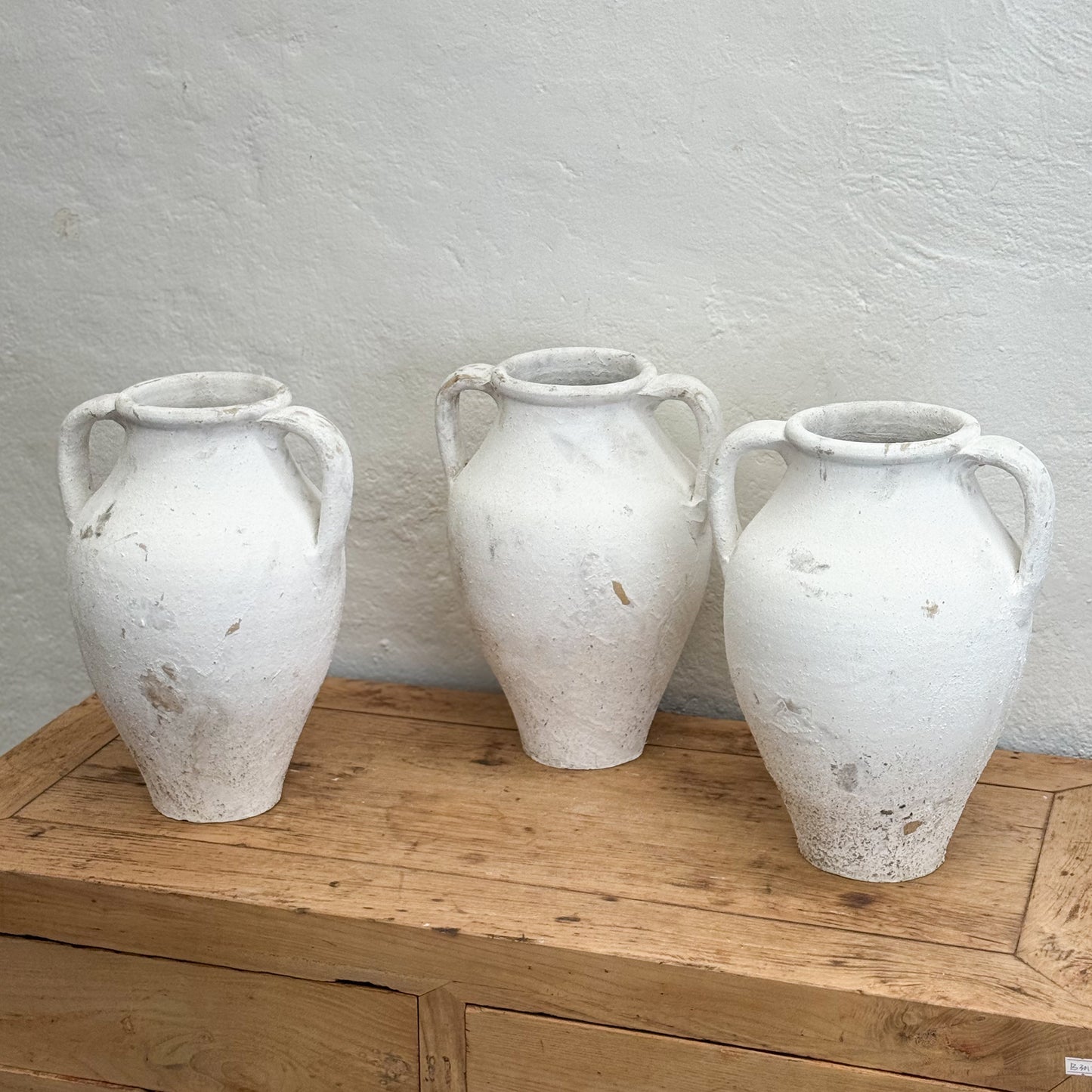 Three white ceramic vases with handles on a wooden surface against a light gray wall.