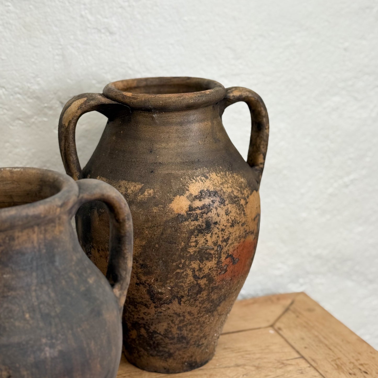 Two rustic clay pots with handles on a wooden surface against a textured white wall.