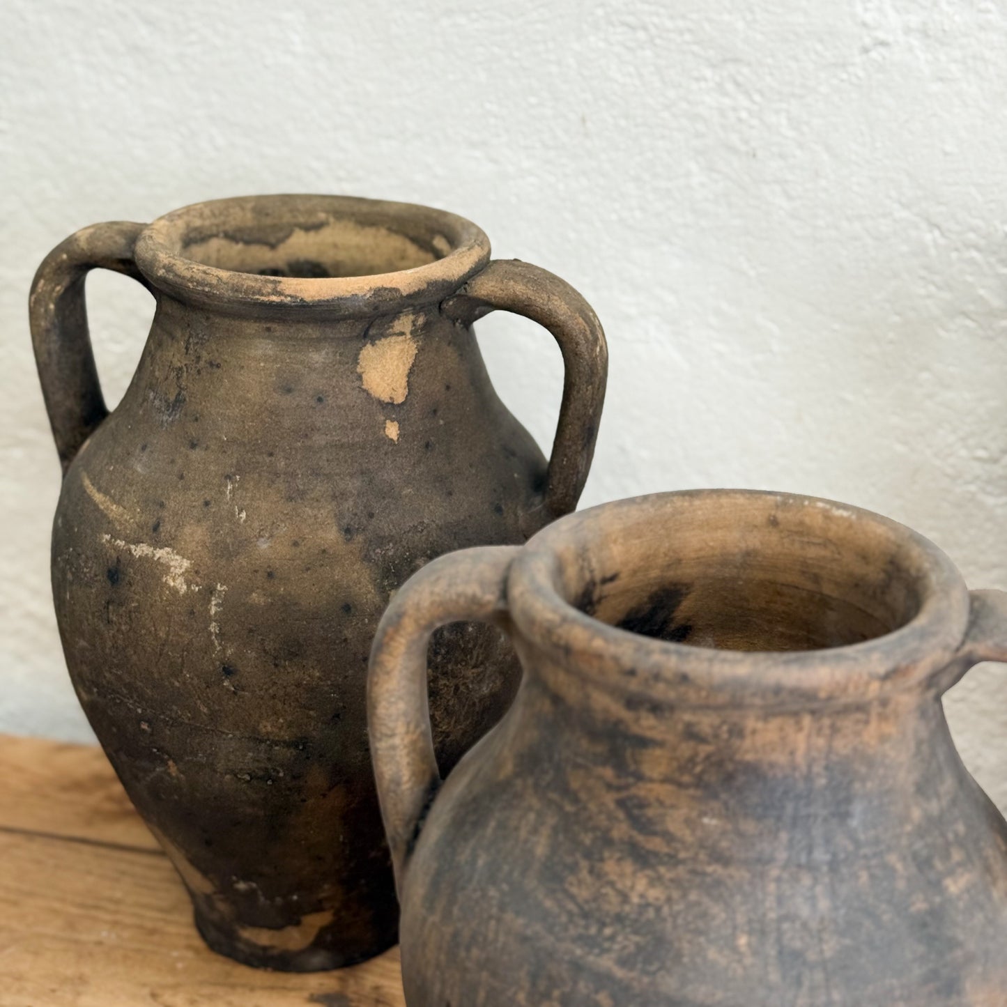 Two rustic clay pots with handles on a wooden surface against a white textured wall.