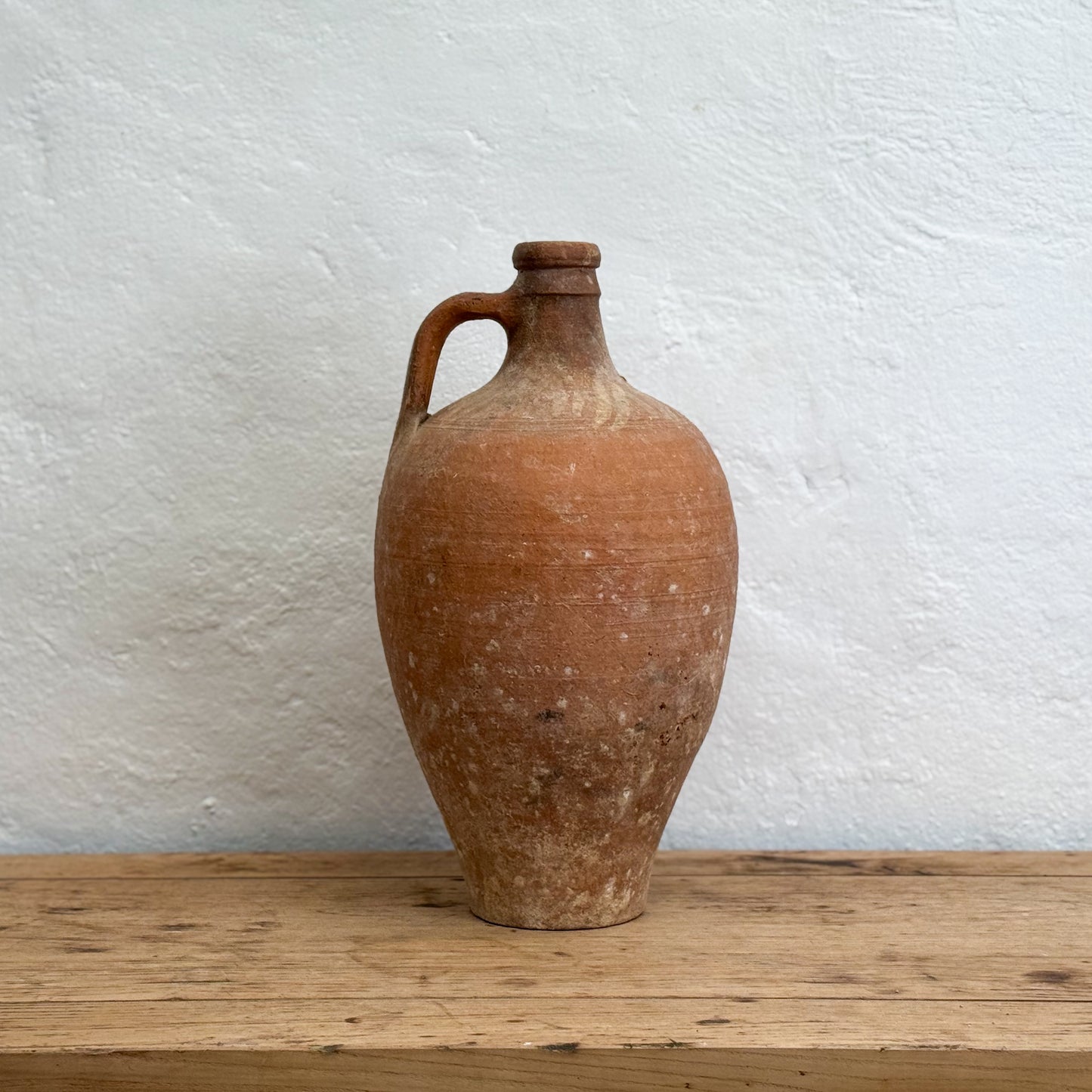 Terracotta jar with a handle on a wooden surface against a textured white wall