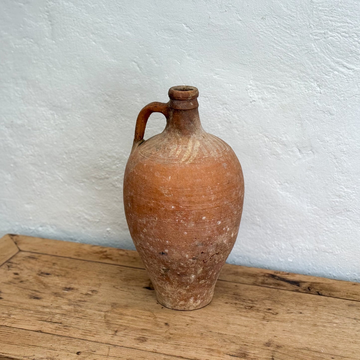 Terracotta jar with a handle on a wooden surface against a white wall