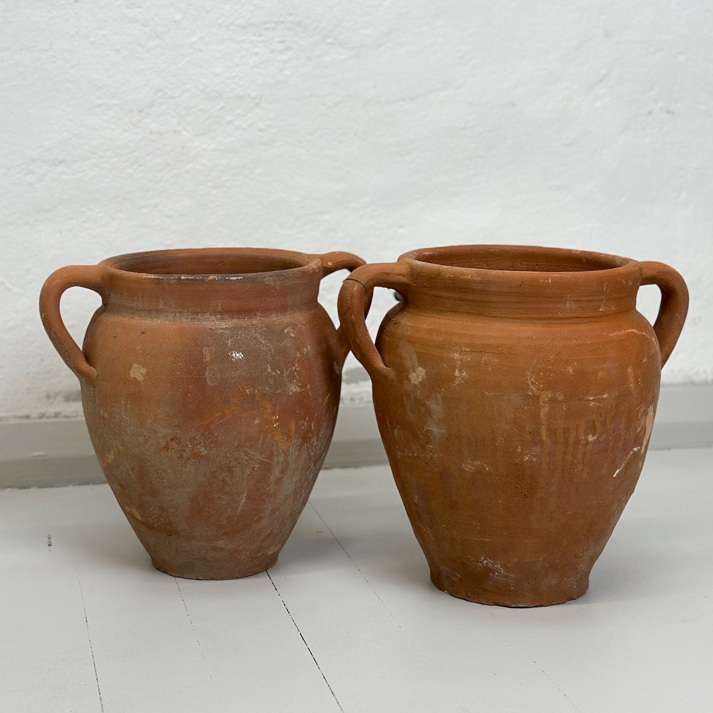 Two brown terracotta pots with handles, placed on a white surface against a white wall.