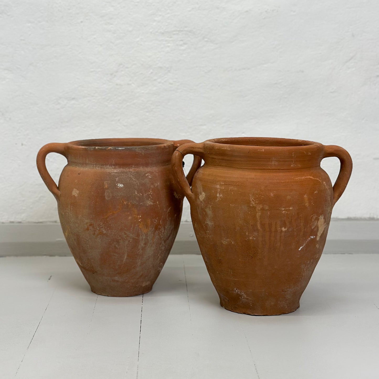 Two brown terracotta pots with handles, placed on a white surface against a white wall.