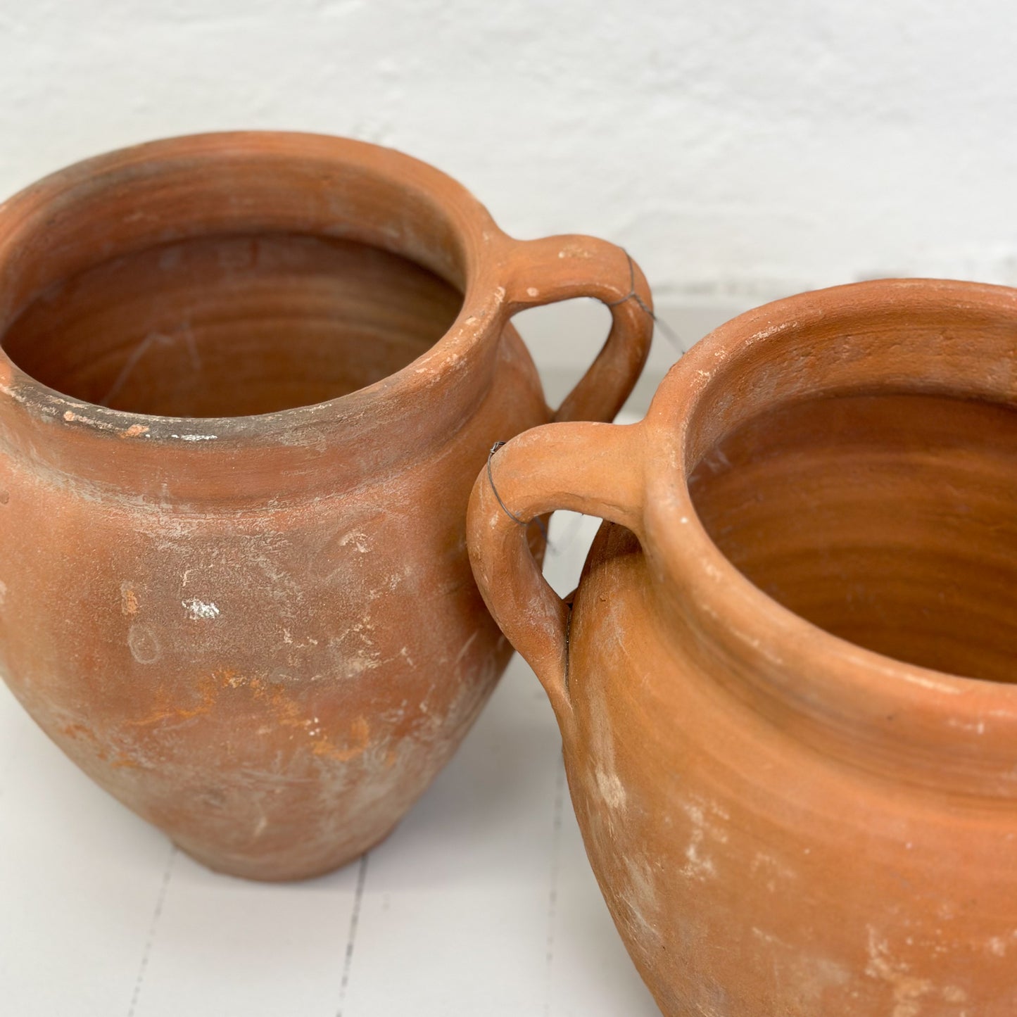 Two brown terracotta pots with handles, placed on a white surface against a white wall.