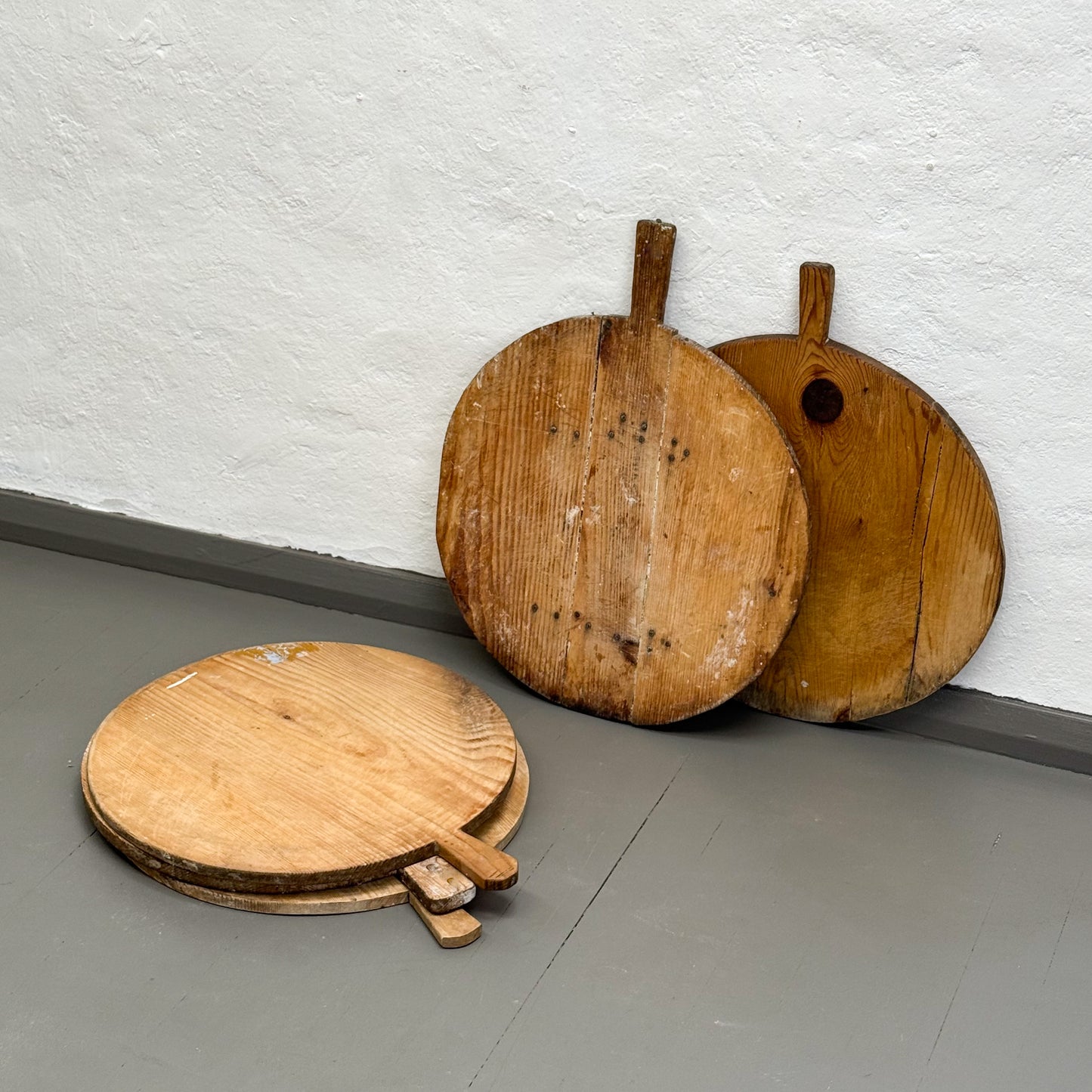 Three wooden pizza peel tools leaning against a white wall on a gray floor.