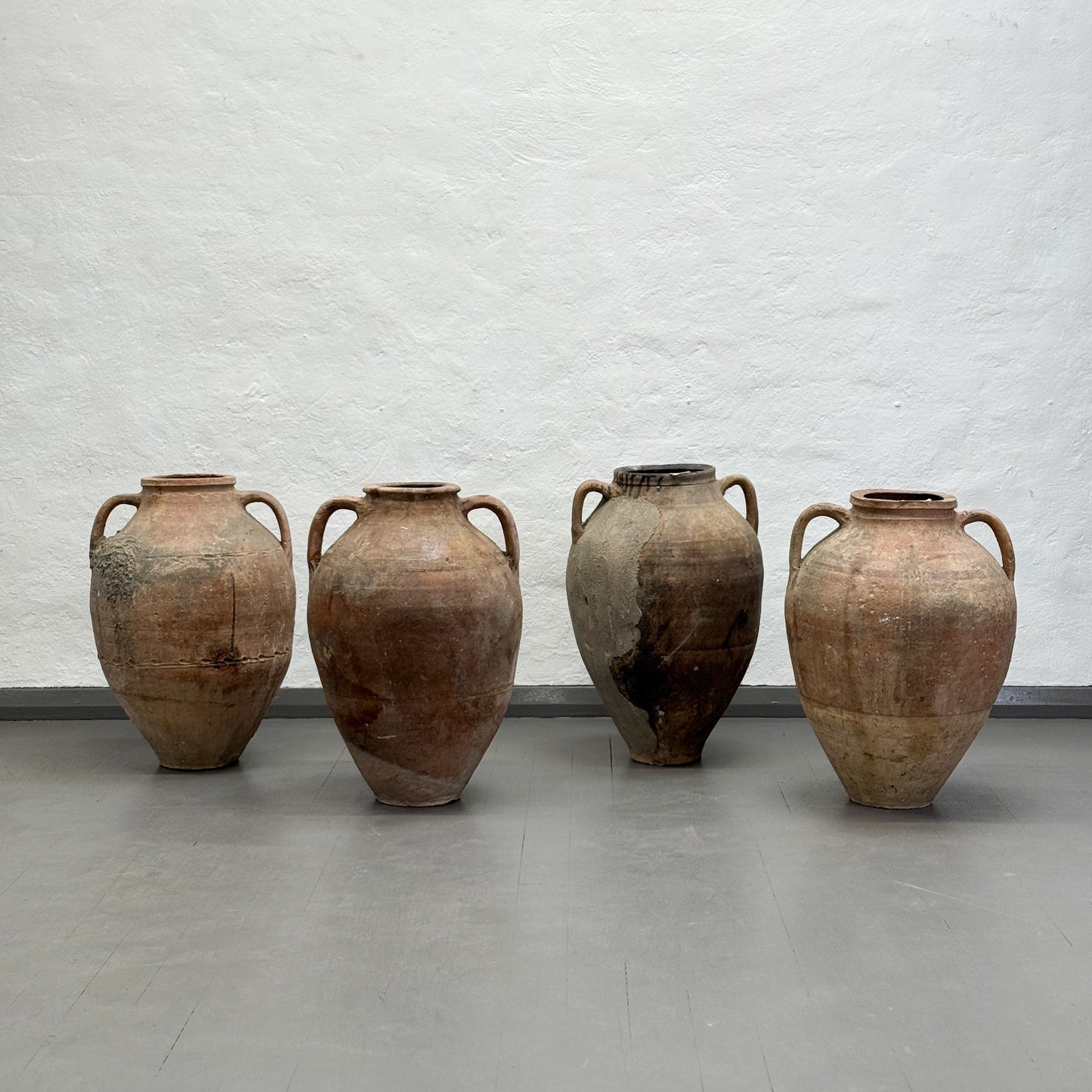 Four ancient-looking clay pots with handles on a gray floor against a light gray textured wall.