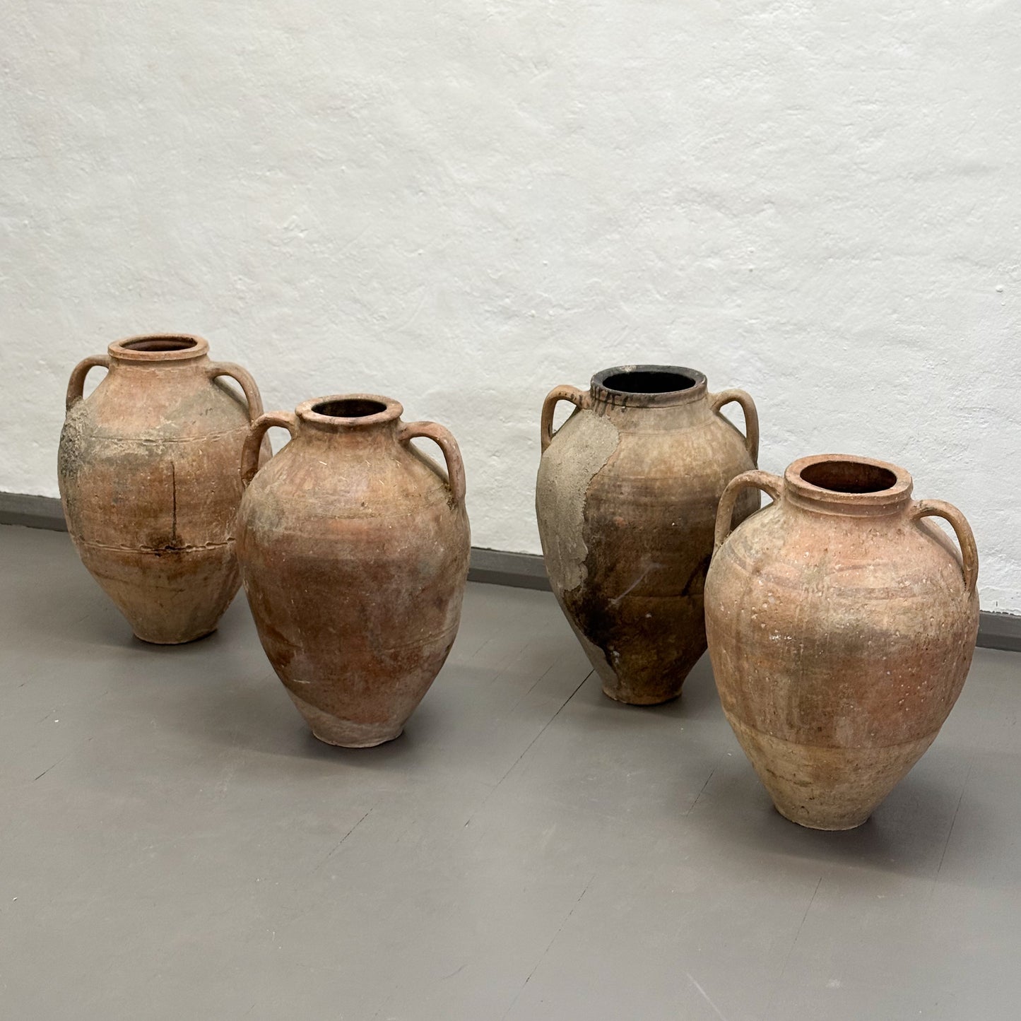 Four large terracotta pots with handles on a gray floor against a light gray wall.