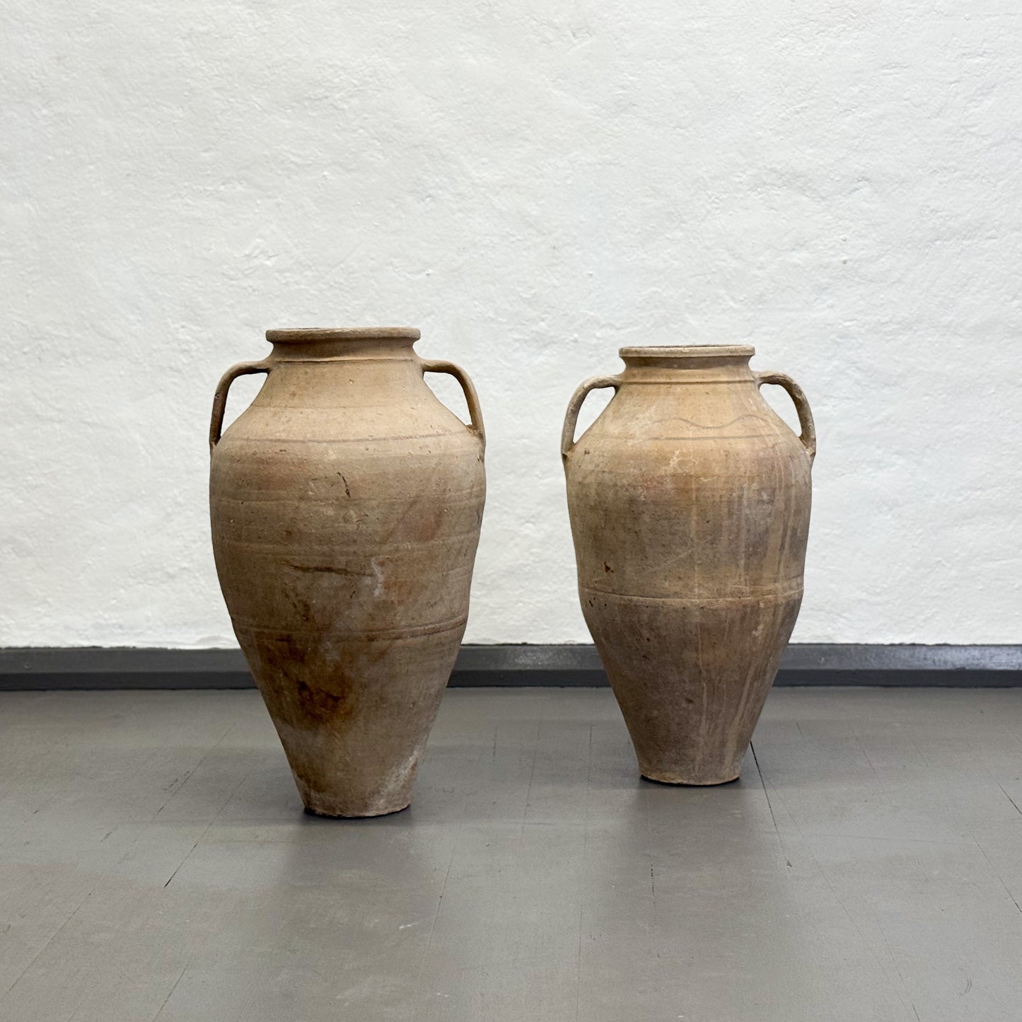 Two large beige urn-shaped vases on a gray floor with a white textured wall background.
