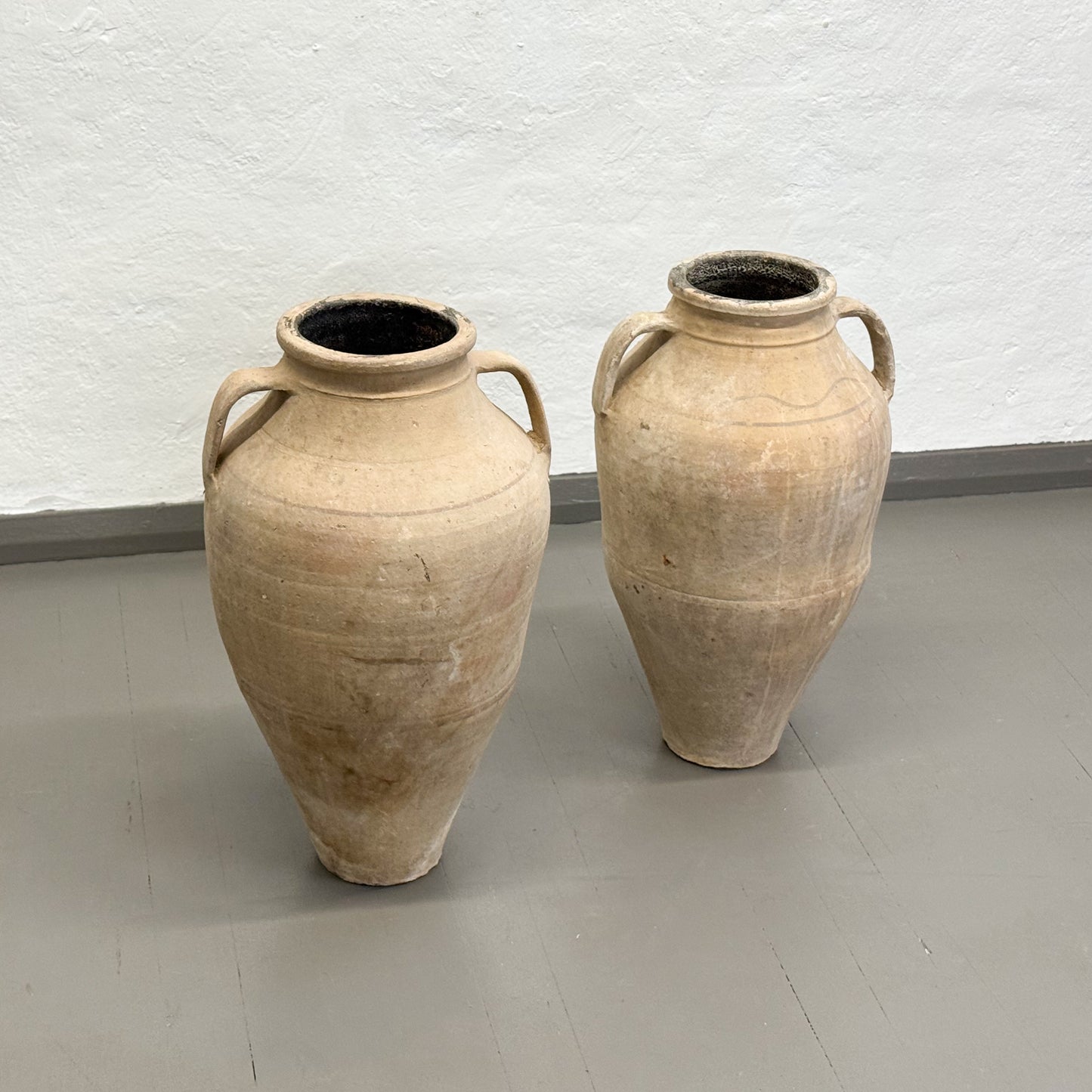 Two large beige ceramic vases with handles on a plain floor.