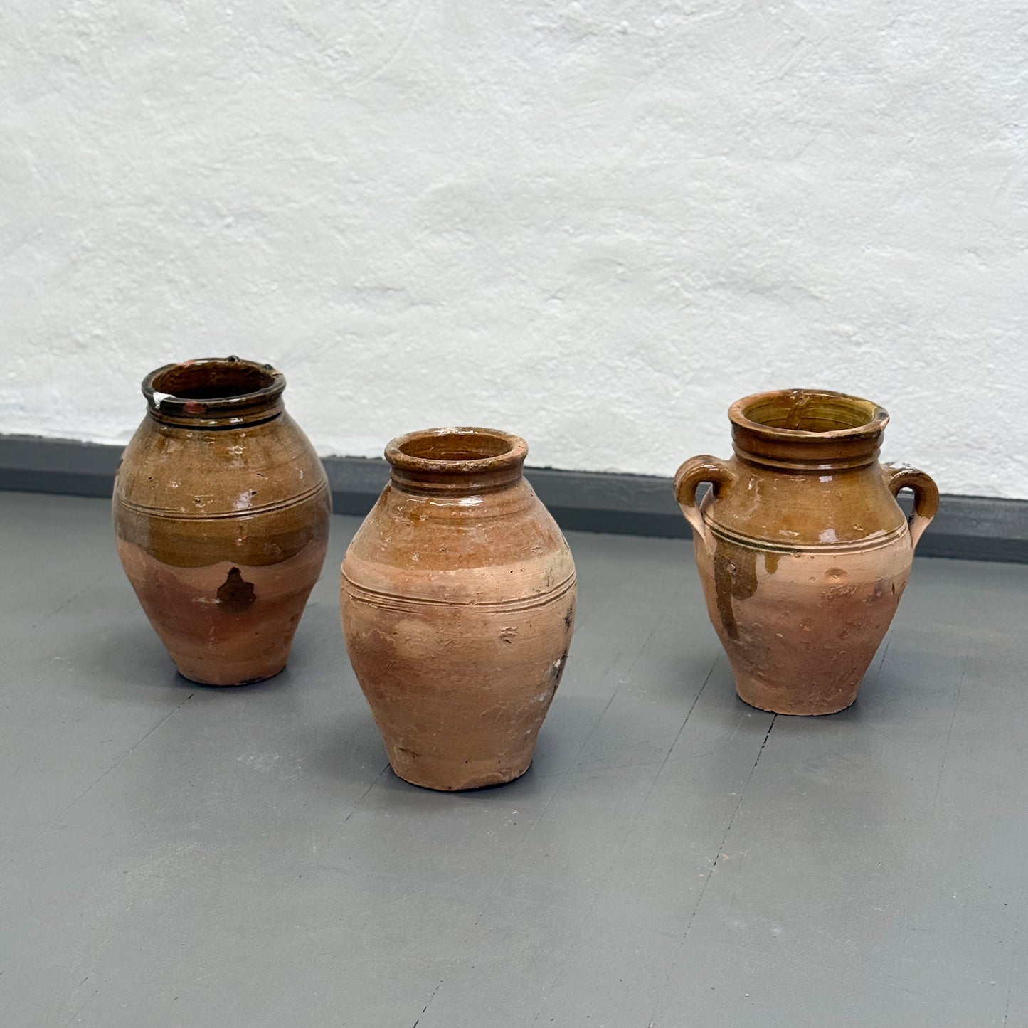 Three ceramic jars of varying sizes on a gray floor with a textured white wall background.