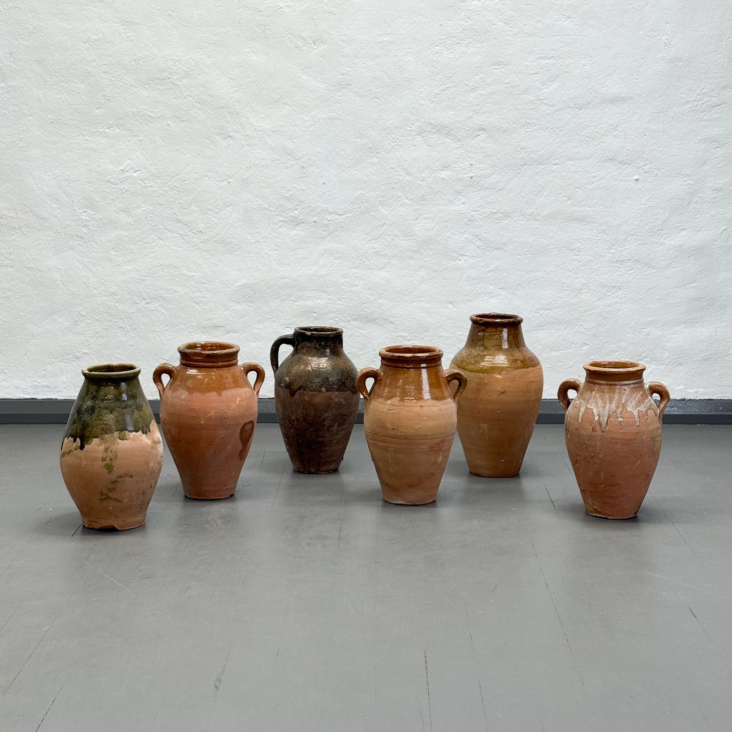 Set of six ceramic vases on a gray floor against a white textured wall.