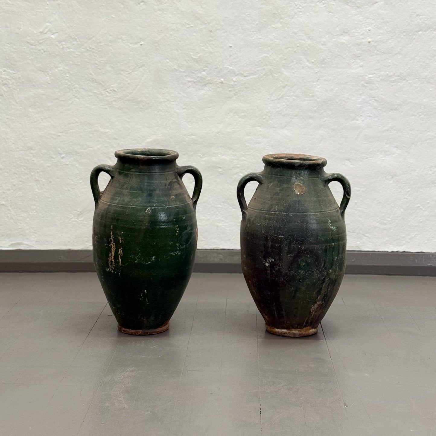 Two green ceramic vases with handles on a plain floor against a white wall.