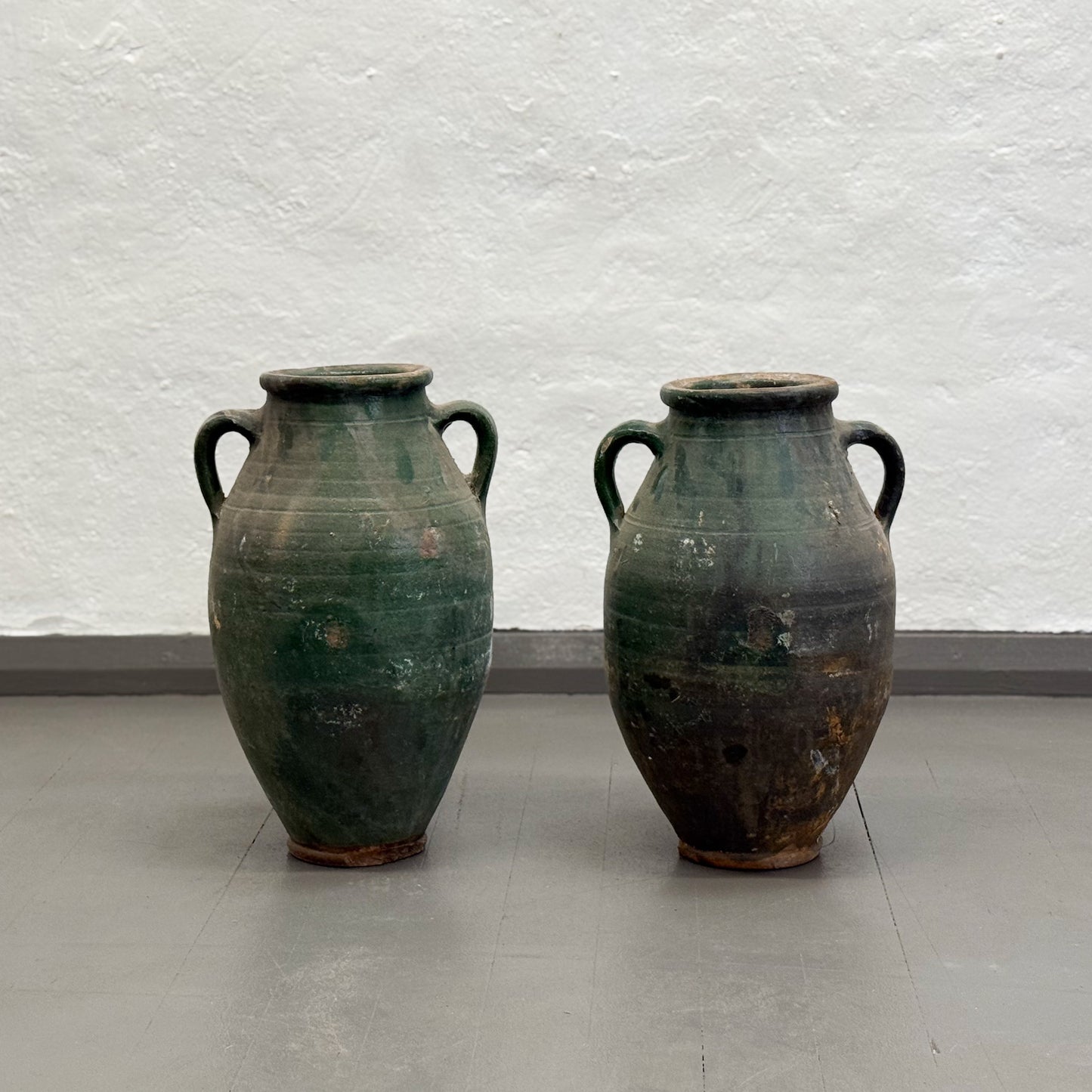 Two green ceramic vases with handles on a plain floor against a white wall.