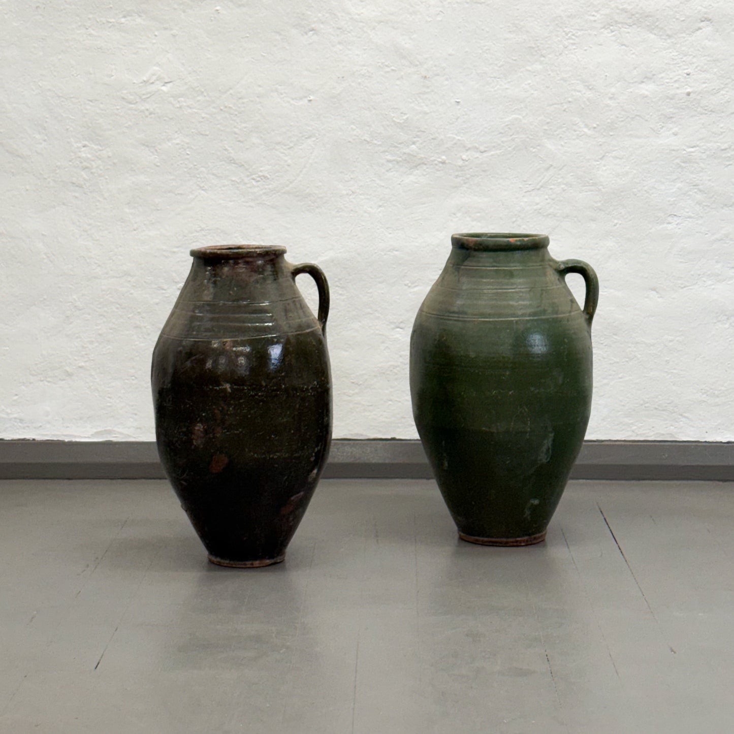 Two green ceramic vases on a plain floor with a white textured wall background
