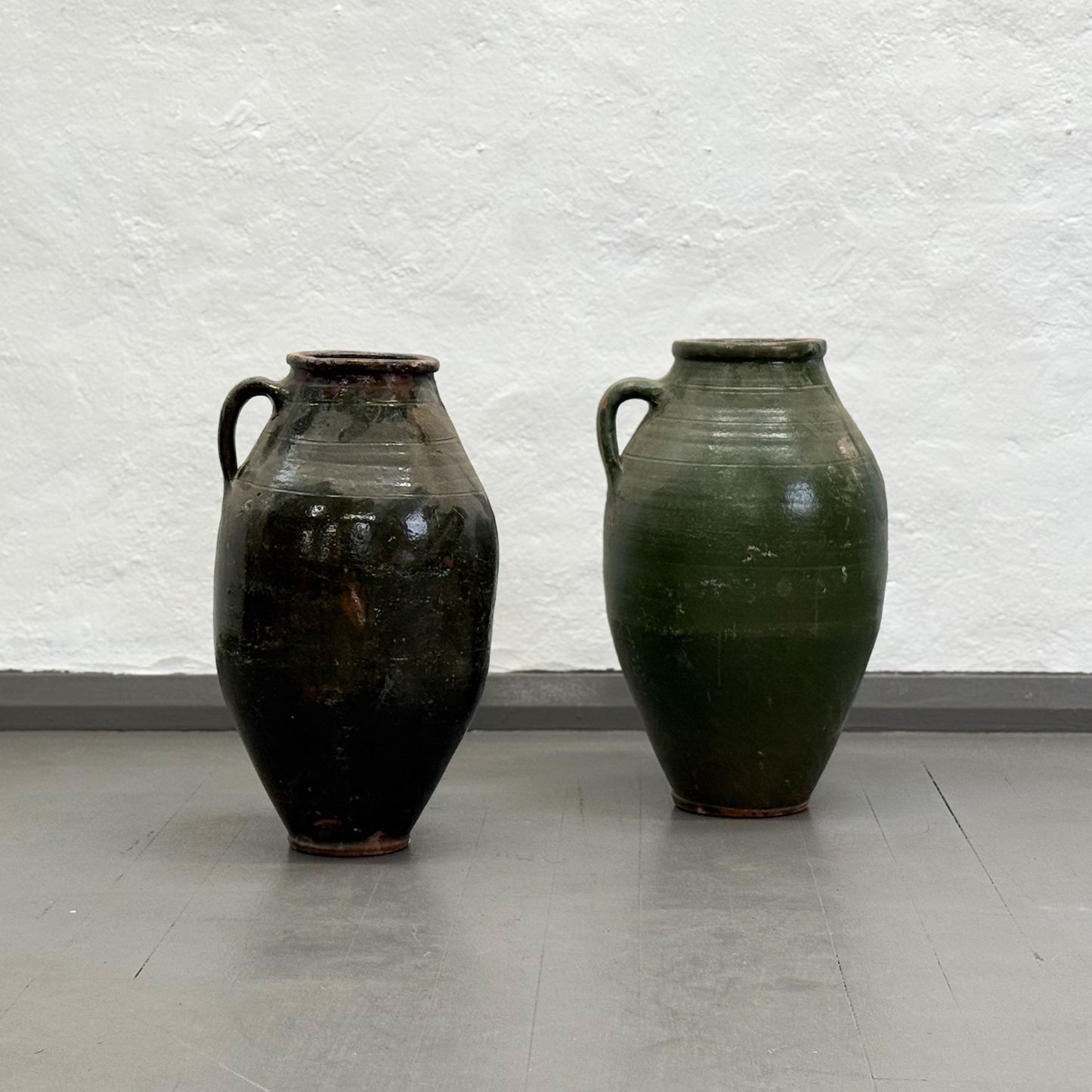 Two green ceramic vases on a gray floor with a textured white wall background